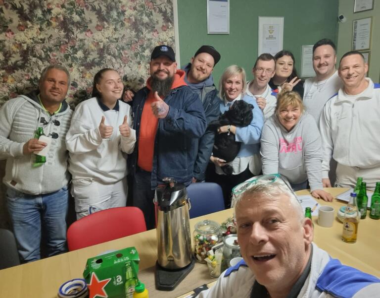 Gruppe von Erwachsenen posiert für ein Gruppen-Selfie; vorderste Person hält die Kamera. Im Raum Tisch mit Thermoskanne, Getränkeflaschen und Süßigkeitenglas; eine Frau hält einen kleinen schwarzen Hund.