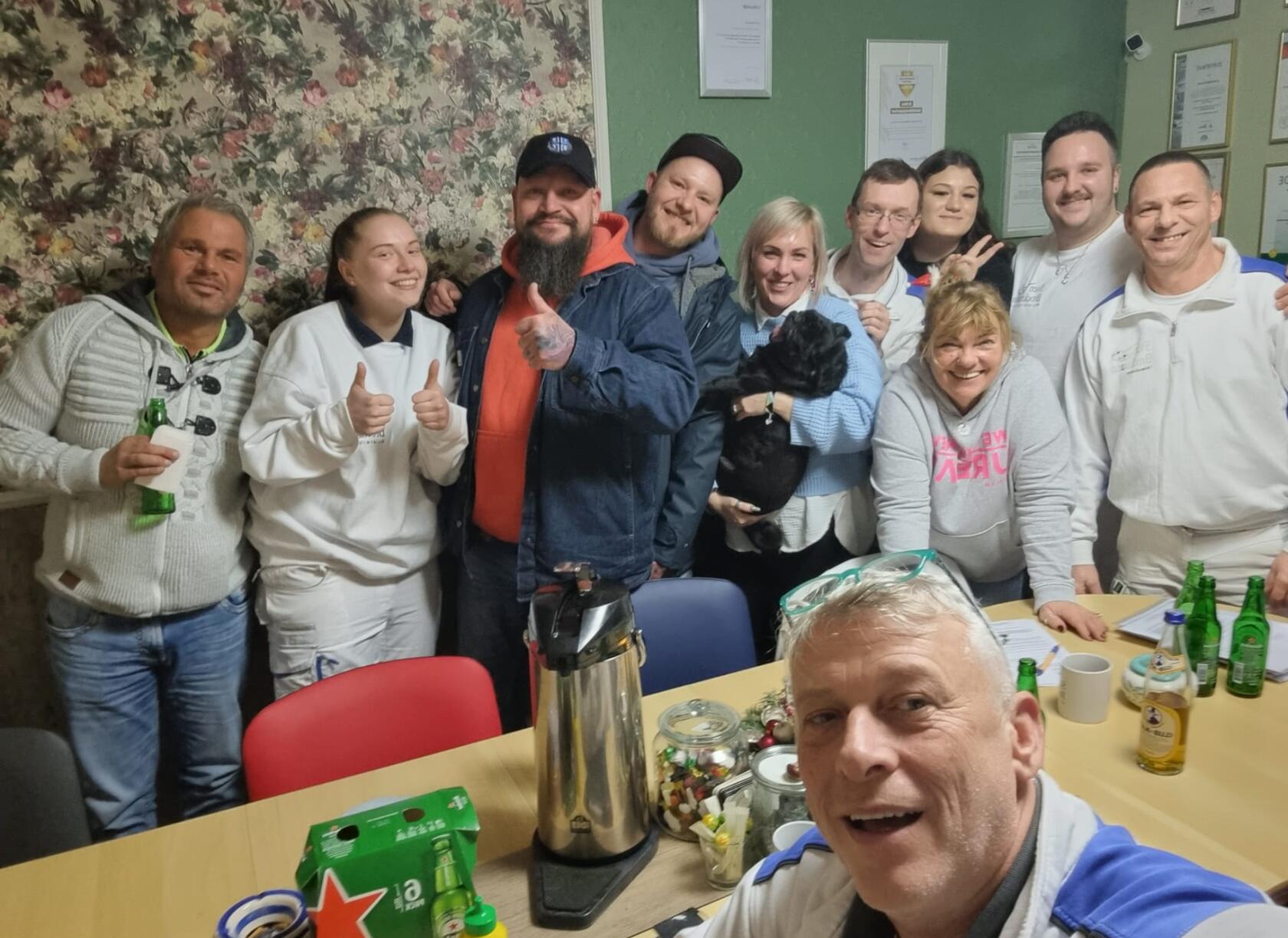 Zwölf Personen posieren dicht beieinander für ein Selfie; eine Person hält die Kamera im Vordergrund. Raum mit Tisch, Kaffeekanne, Flaschen, Süßigkeiten und floralem Tapetenmuster.