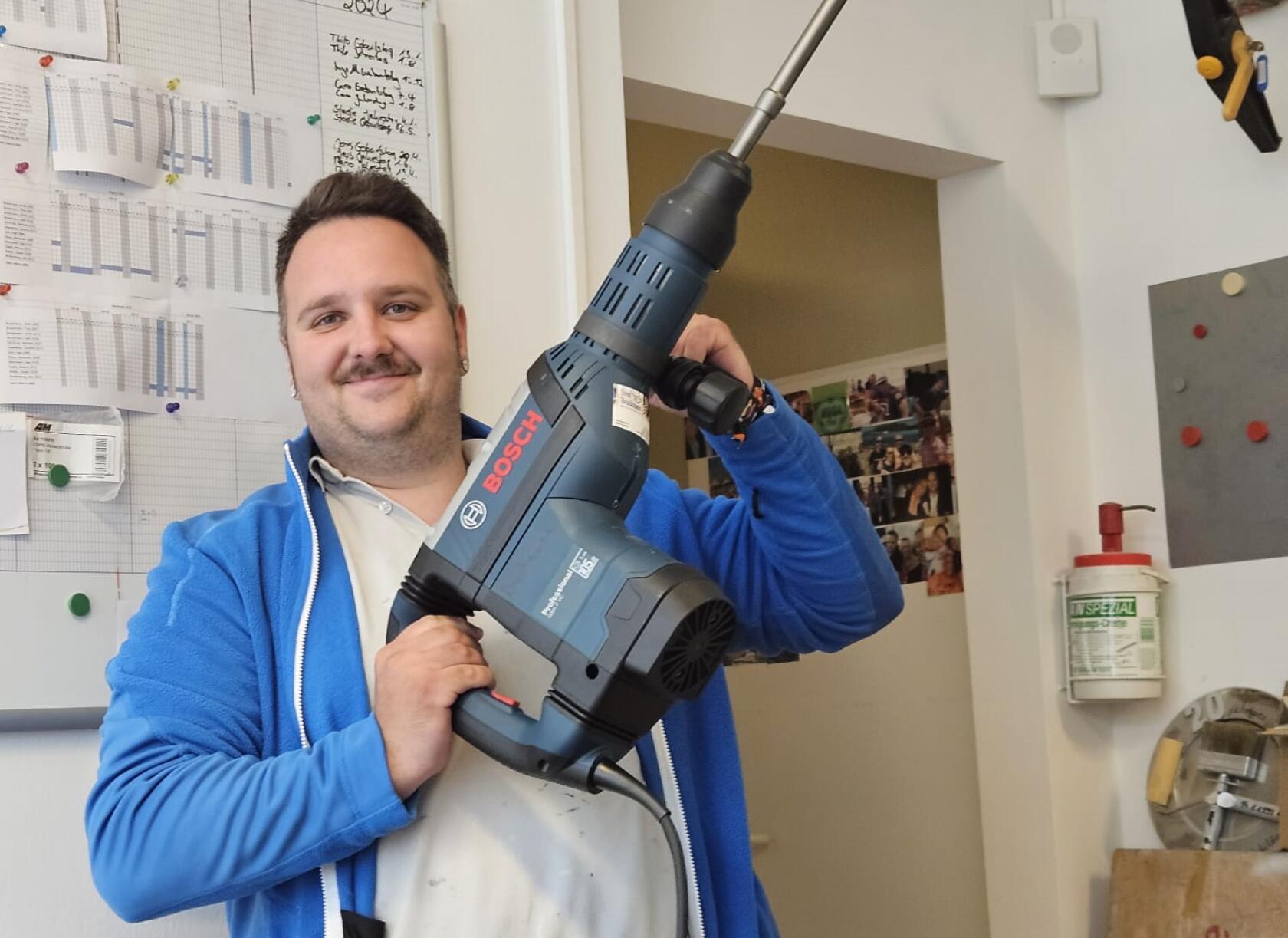 Mann hält großen blauen BOSCH-Bohrhammer schräg vor dem Körper; Hintergrund: Pinnwand mit Tabellen und handschriftlicher Notiz 