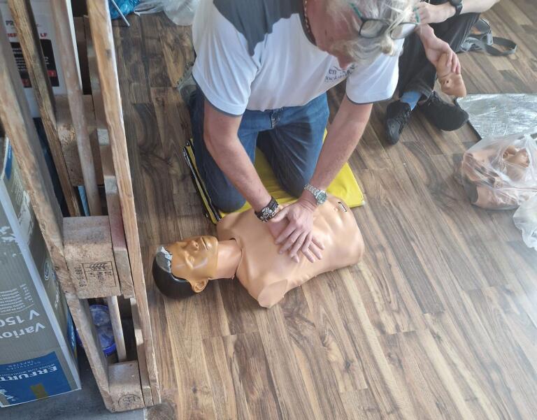 Erwachsener kniet und führt Herzdruckmassage an halbem Torso-Trainingsdummy aus, auf Holzboden mit gelber Knieunterlage; weitere Puppe, Person und Palettenkartons im Raum sichtbar.