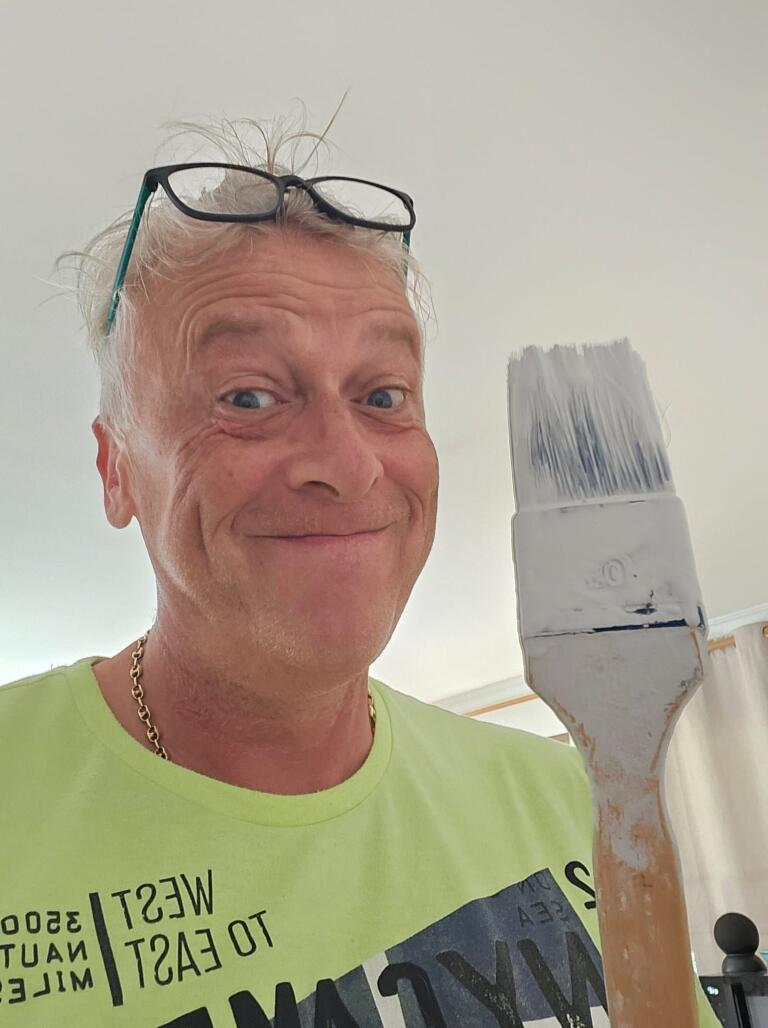 Mann hält großen Flachpinsel mit weißer Farbe nahe an die Kamera, blickt in die Kamera; Brille auf dem Kopf, trägt hellgrünes T‑Shirt mit Aufdruck 