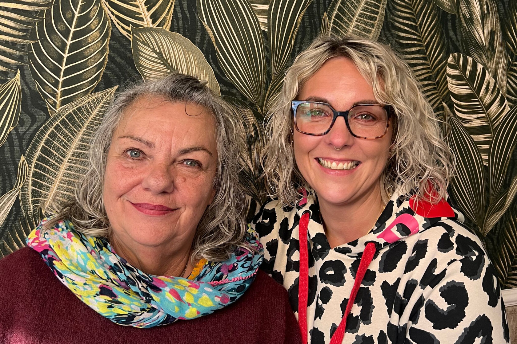 Zwei lächelnde Frauen, links mit grauem Haar und buntem Schal, rechts mit lockigem blondem Haar und Brille in Leopardenmuster-Hoodie; goldene Blatt-Tapete im Hintergrund.