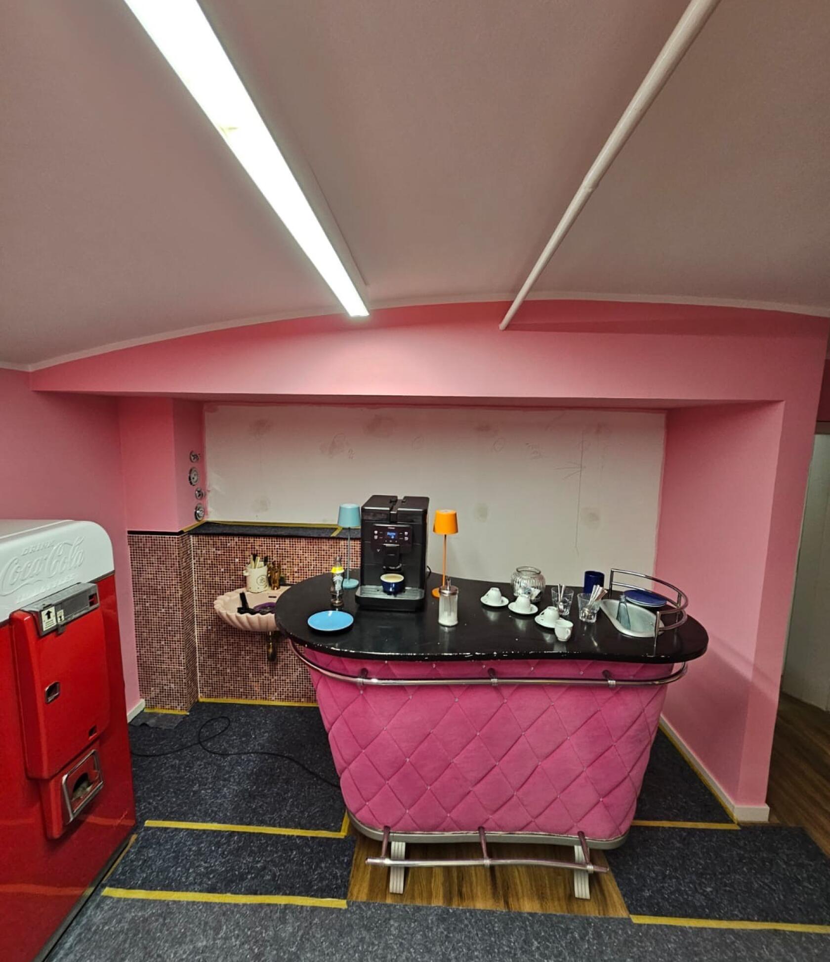 Rosa gestepptes Barpult steht zentral, darauf Kaffeemaschine, Tassen und kleine Lampen; kleiner Raum mit rosa Wänden, Deckenleuchte oben und rotem Kühlschrank links (Coca-Cola).