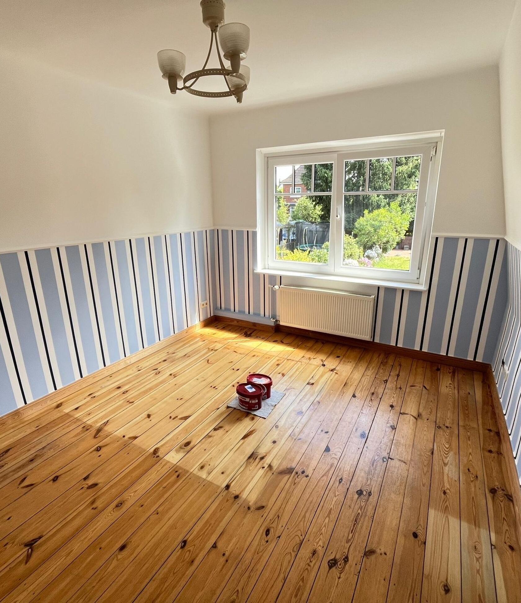 Zimmer mit halbhoher blau-weiß gestreifter Tapete — Sonnenlicht fällt durch das Fenster auf den Holzdielenboden; zwei Farbdosen auf Boden, Heizkörper und Deckenlampe sichtbar.
