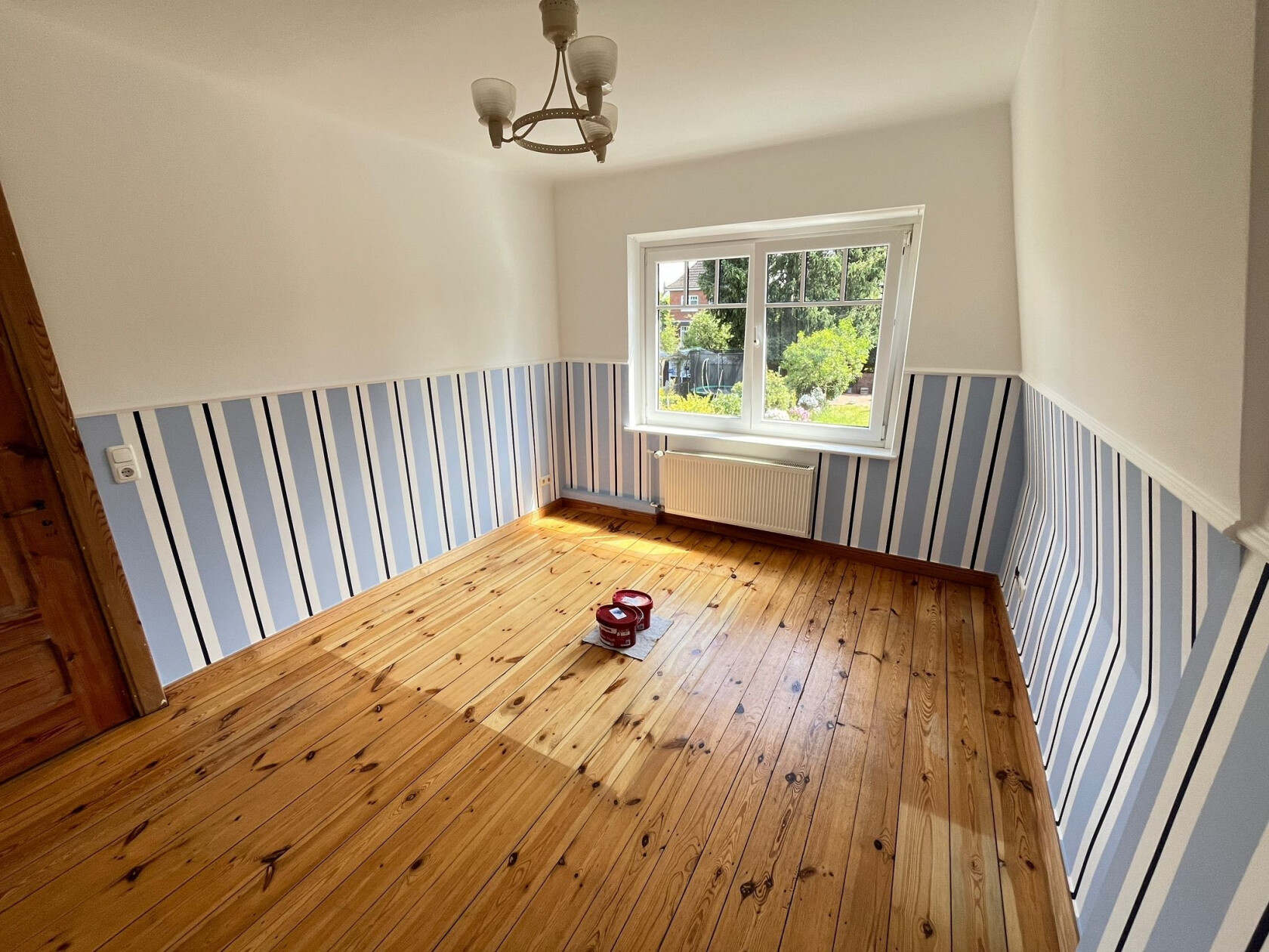 Zimmer mit halbhoher blau-weiß gestreifter Tapete — Sonnenlicht fällt durch das Fenster auf den Holzdielenboden; zwei Farbdosen auf Boden, Heizkörper und Deckenlampe sichtbar.