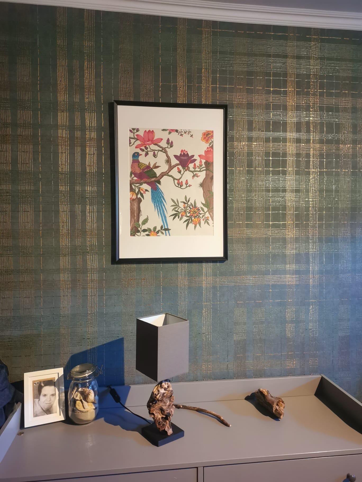 Gerahmtes Bild mit farbigem Vogel auf Ast hängt an dunkelgrüner Tapete mit goldenen Linien; darunter grauer Schreibtisch mit grauem Lampenschirm, Glasbehälter mit Muscheln, gerahmtes Schwarzweißporträt und Treibholz.