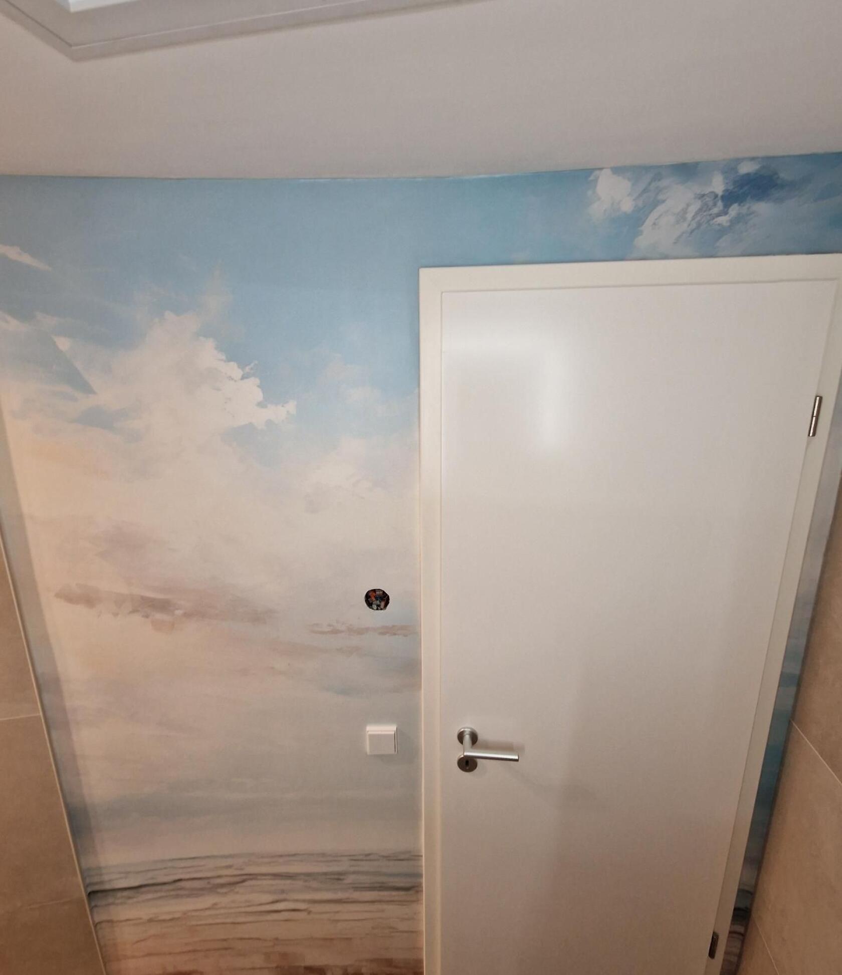 Weiße geschlossene Tür mit silbernem Griff in einer Wand mit großflächigem Himmel- und Strandwandbild; links sichtbarer Lichtschalter und runde Öffnung für Steckdose.