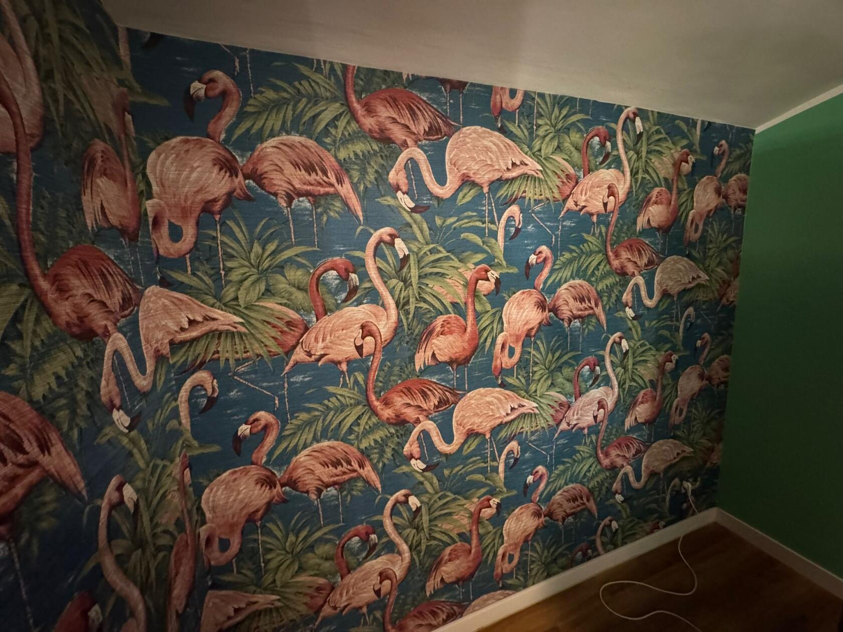 Tapete mit zahlreichen rosa Flamingos, die teilweise stehen und die Hälse neigen, auf blauem Hintergrund mit grünen Palmblättern; rechter Wandrand grün, Holzfußboden und weißer Sockel sichtbar.