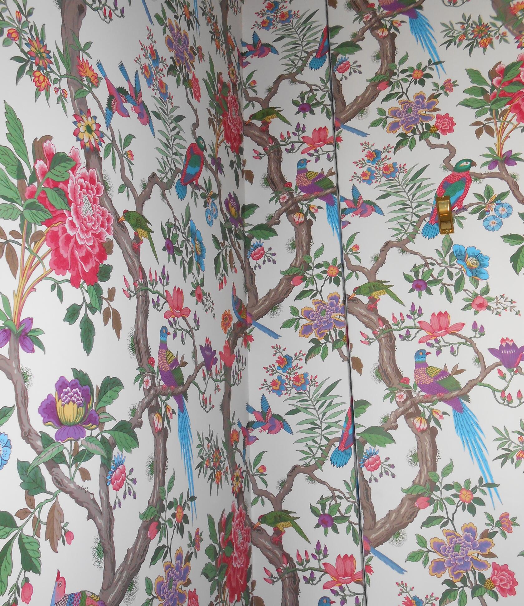 Tapetenmuster mit bunten Vögeln, einige sitzend und einige im Flug, auf verschlungenen Baumzweigen mit großen Blumen; weiße Hintergrundfläche, sichtbare Raumecke und goldener Lichtschalter.