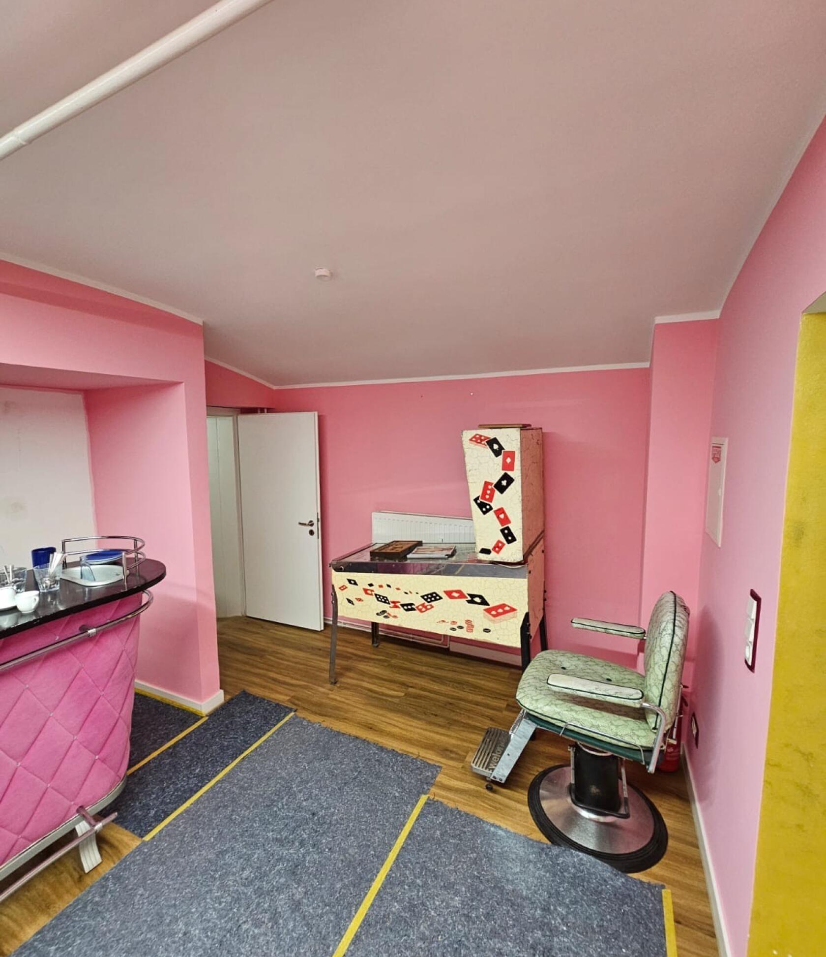 Hellgrüner Friseurstuhl auf schwarzer Metallbasis steht nahe einer Wand in einem pink gestrichenen Raum mit Holzfußboden, dekoriertem Flipperautomaten, rosa Theke, geöffneter weißer Tür und grauen Abdeckmatten.