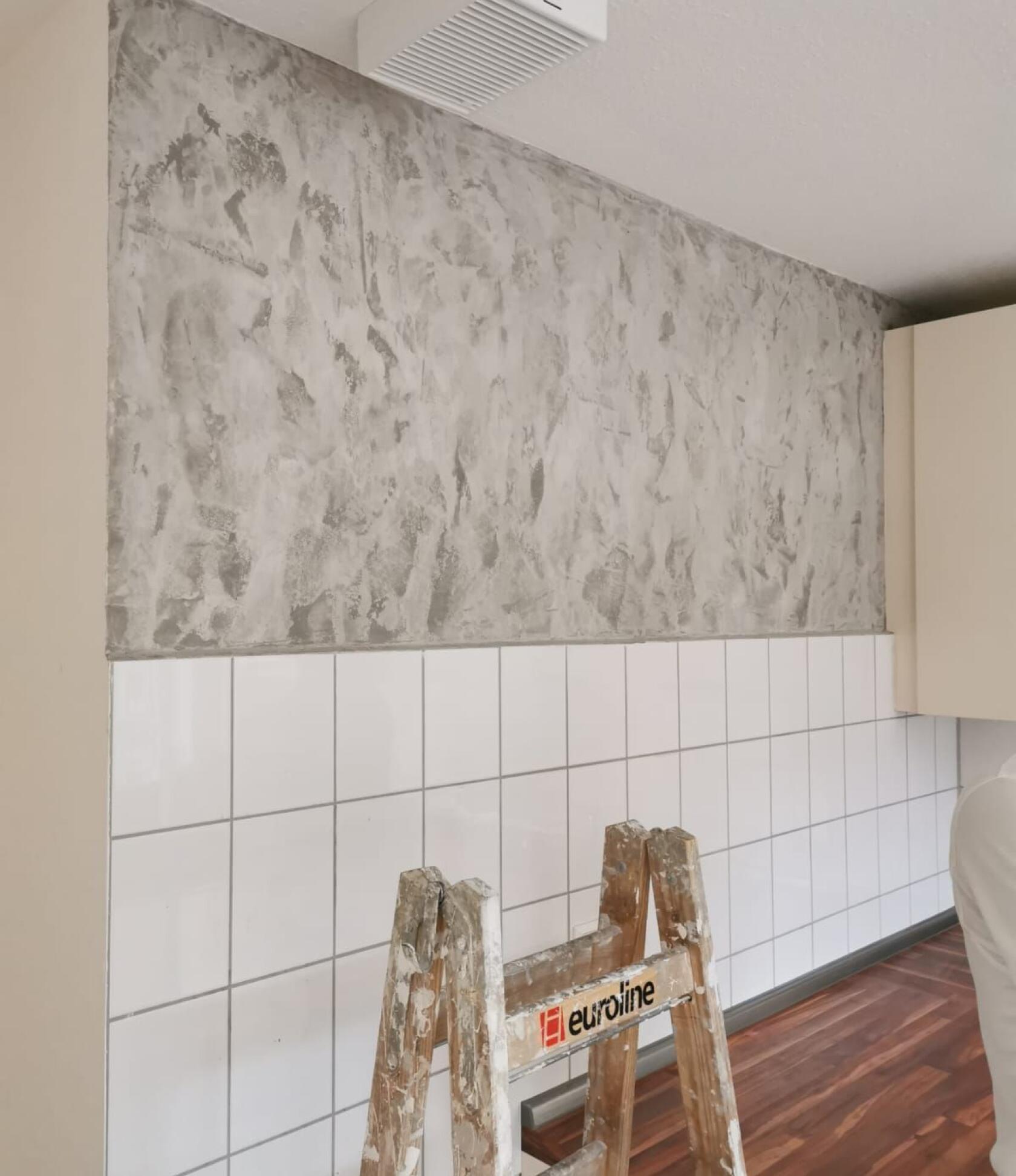 Holzleiter mit Farbspritzern und Aufdruck „euroline“ steht vor weißen quadratischen Fliesen; darüber grau strukturiert verputzte Wand, rechts Küchenschrank und brauner Holzboden sichtbar.