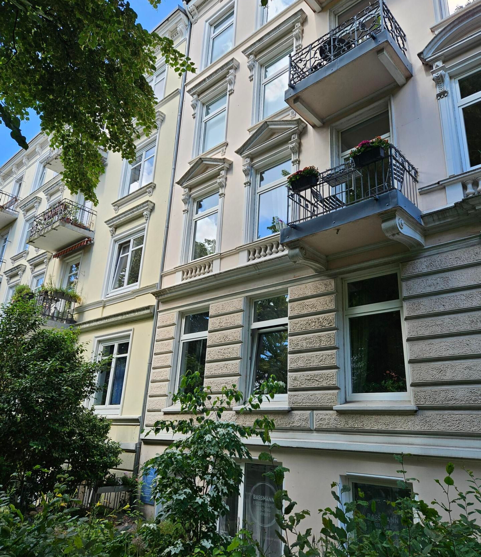 Mehrstöckige Altbaufassade in vier Stockwerken trägt schmiedeeiserne Balkone mit Blumenkästen – Vorgarten mit Sträuchern und Bäumen, weiße Fenster mit Stuckrahmen und sandfarbener Putzstruktur.