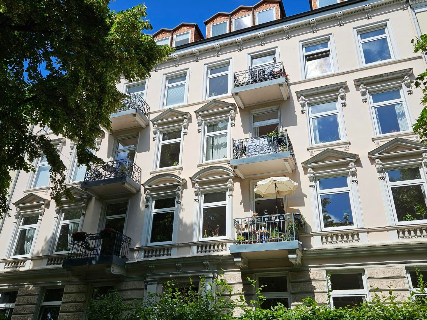 Fassade eines vierstöckigen Altbaus zeigt drei Balkone mit Pflanzen und offenem Sonnenschirm, weiße Fensterrahmen, verzierte Gesimse, hellbeige Putz, Bäume im Vordergrund.