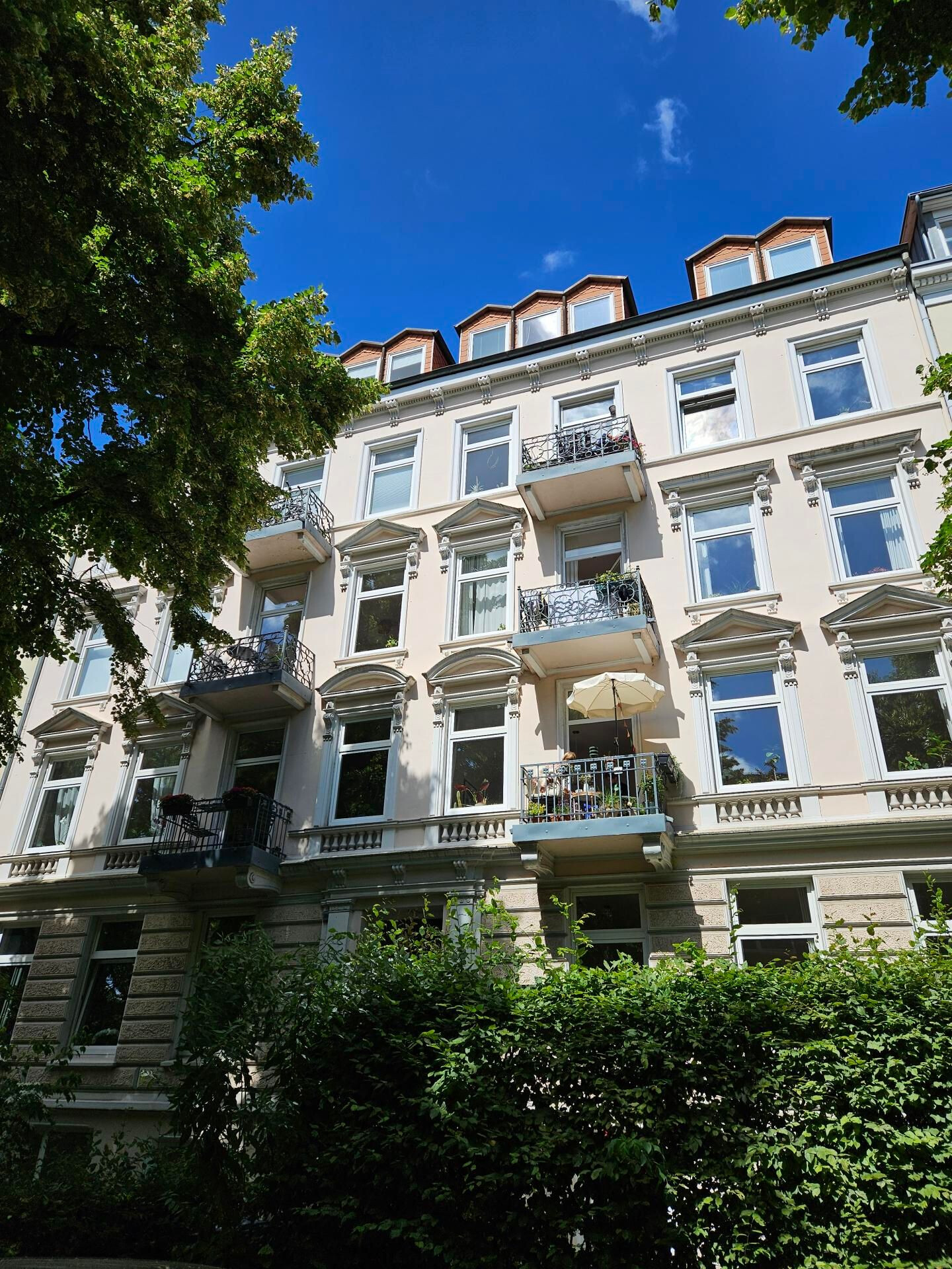 Fassade eines vierstöckigen Altbaus zeigt drei Balkone mit Pflanzen und offenem Sonnenschirm, weiße Fensterrahmen, verzierte Gesimse, hellbeige Putz, Bäume im Vordergrund.
