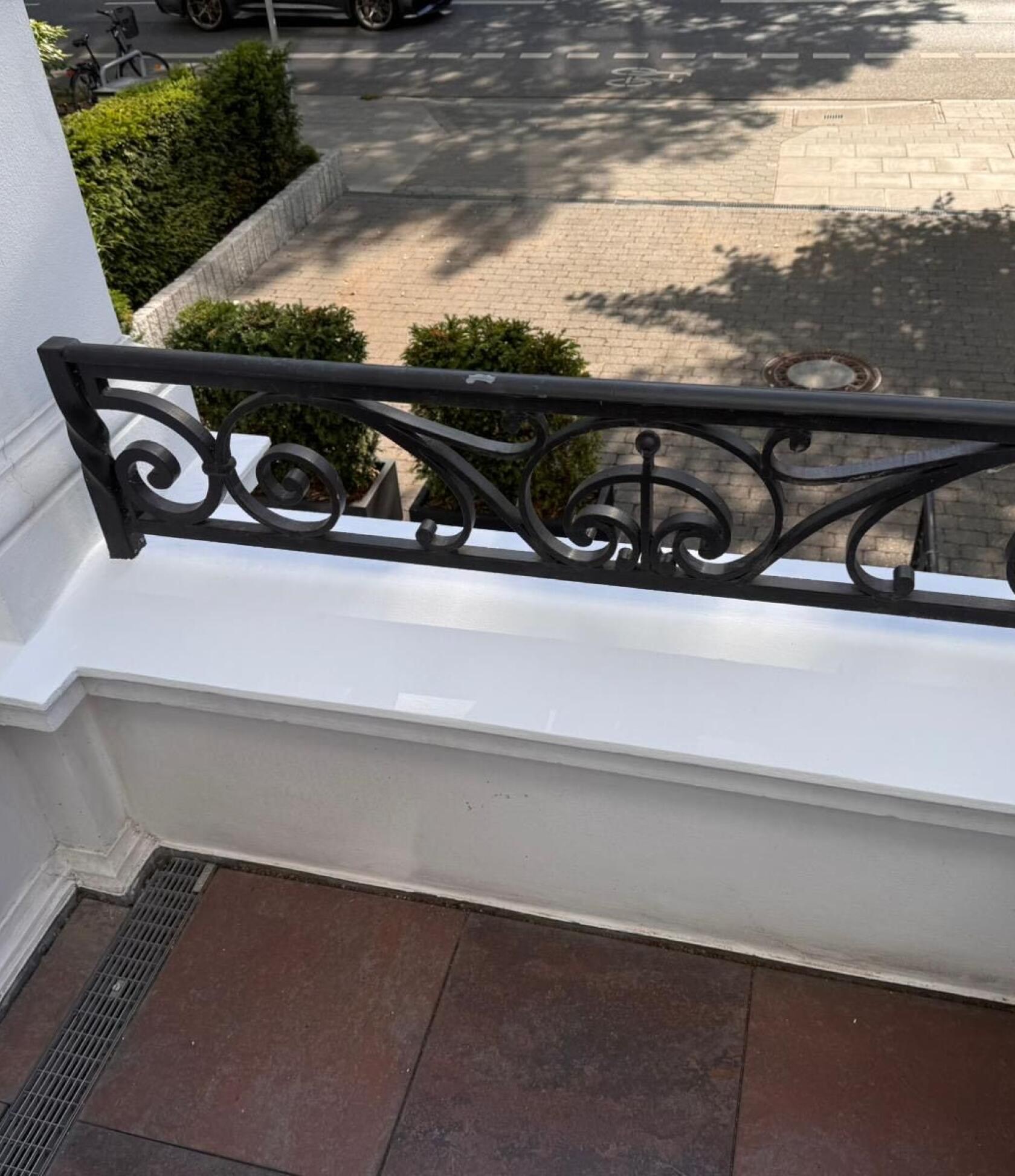 Schmiedeeisernes, schwarzes Balkongeländer mit geschwungenen Ornamenten, montiert auf weißem Balkonsims, begrenzt den Balkon; im Hintergrund gepflasterter Gehweg, niedrige Hecken und Straßenschatten.