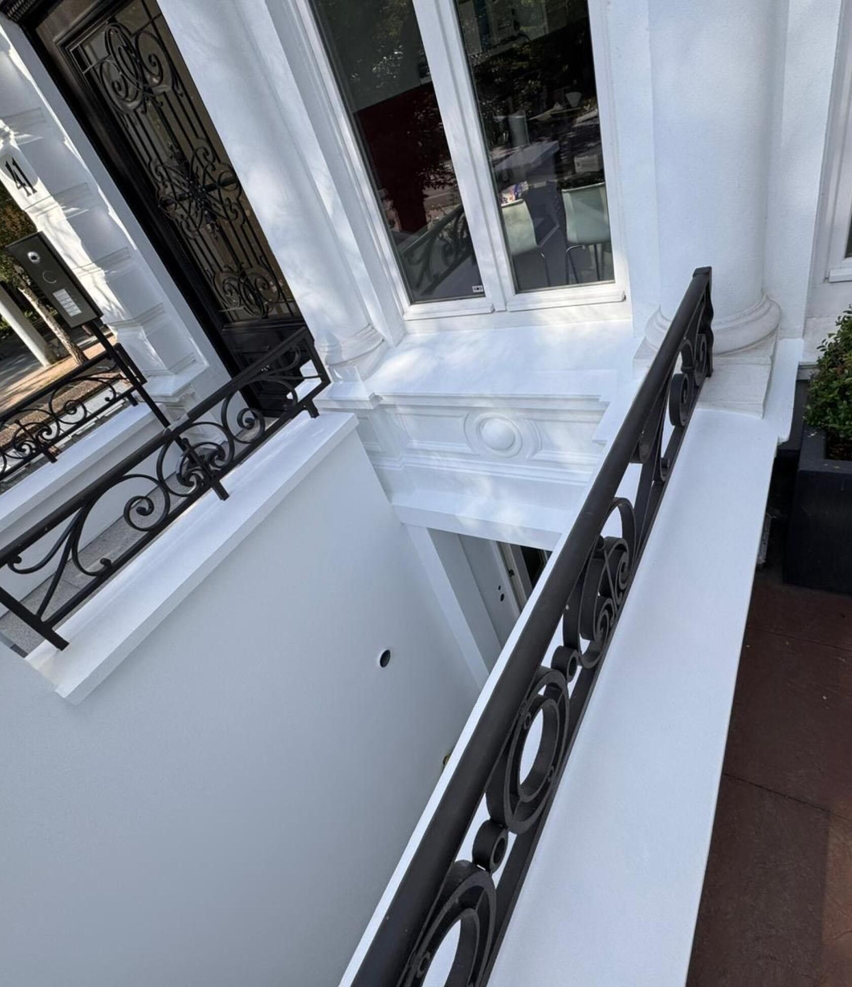 Schwarzes schmiedeeisernes Geländer mit Kreisornamenten verläuft entlang einer weißen Brüstung und überblickt einen hellen Lichthof; weiße Fassadendetails, Fenster mit Inneneinrichtung und schwarze Haustür links.