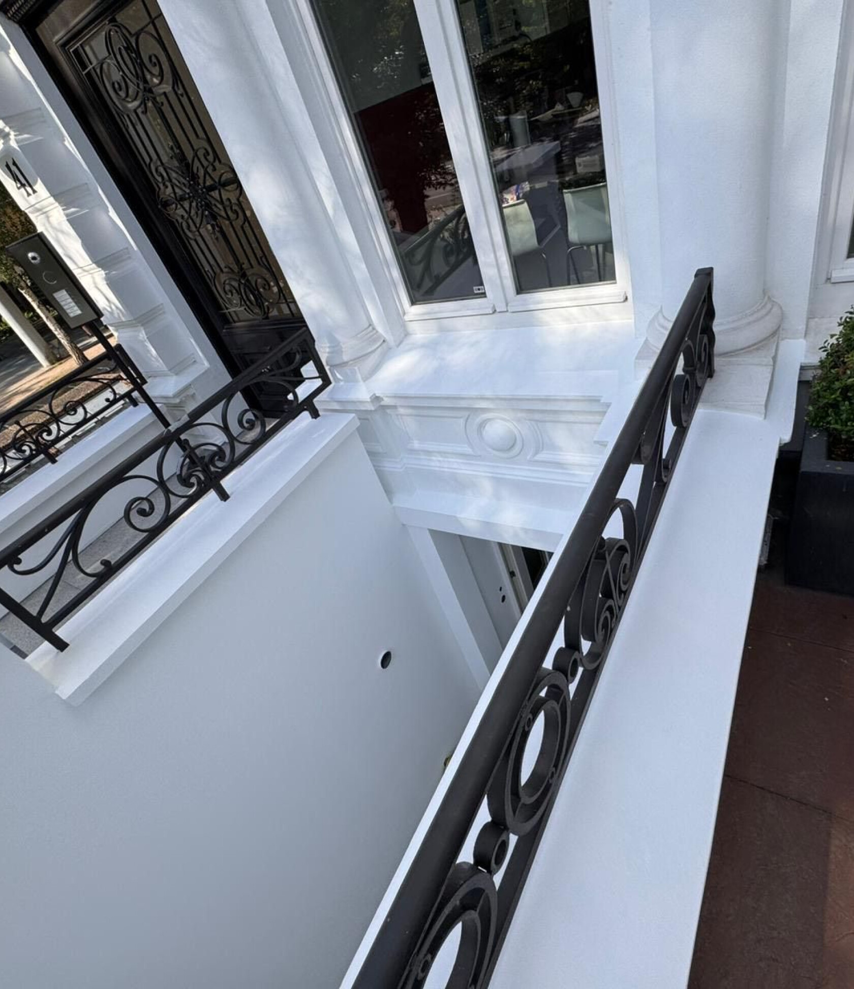 Schwarzes schmiedeeisernes Geländer mit Kreisornamenten verläuft entlang einer weißen Brüstung und überblickt einen hellen Lichthof; weiße Fassadendetails, Fenster mit Inneneinrichtung und schwarze Haustür links.