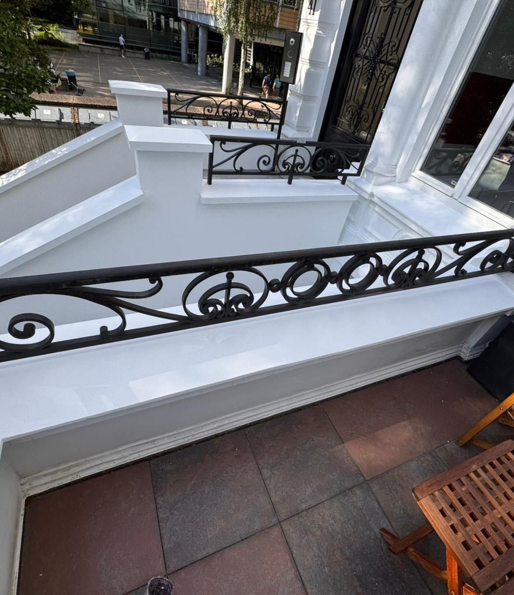 Schwarz geschmiedetes Ziergeländer verläuft entlang einer weißen Balkonbrüstung; Blick auf weiße Fassaden, rote Bodenplatten und einen Holzklappstuhl in der unteren Ecke, Straßenszene im Hintergrund.