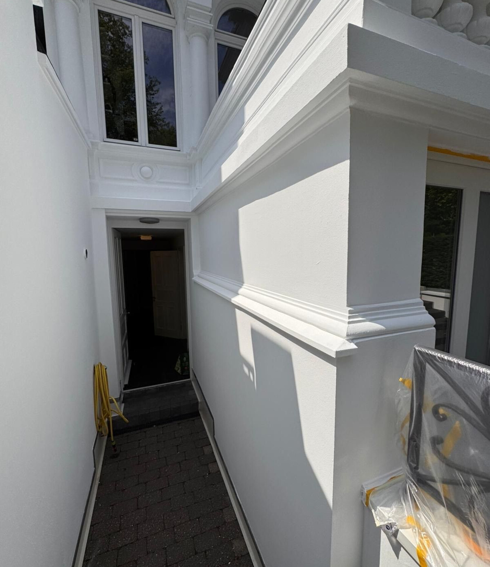 Schmaler weißer Außenflur führt zu offenem dunklem Eingang; Sonnenlicht wirft Schatten auf Wand. Oben dekorative Fenster und Stuck, links aufgerollter gelber Schlauch, rechts plastikabgedecktes Metallgeländer, gepflasterter Boden.