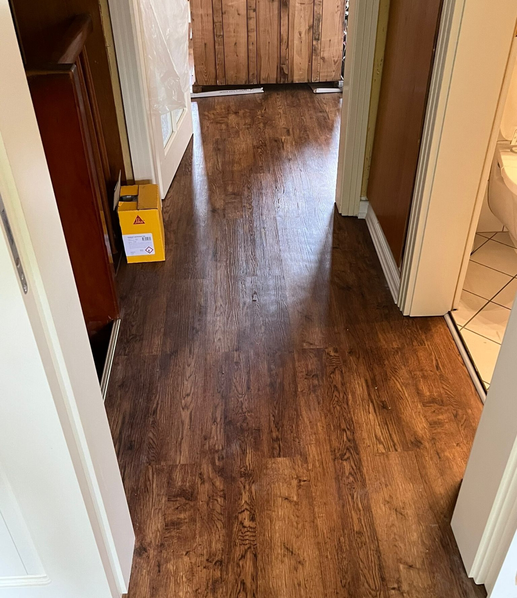 Dunkelbrauner Holzfußboden verläuft durch einen engen Flur mit geöffneten Innentüren rechts und links, gelber Karton neben linker Tür und gefliestes WC rechts.