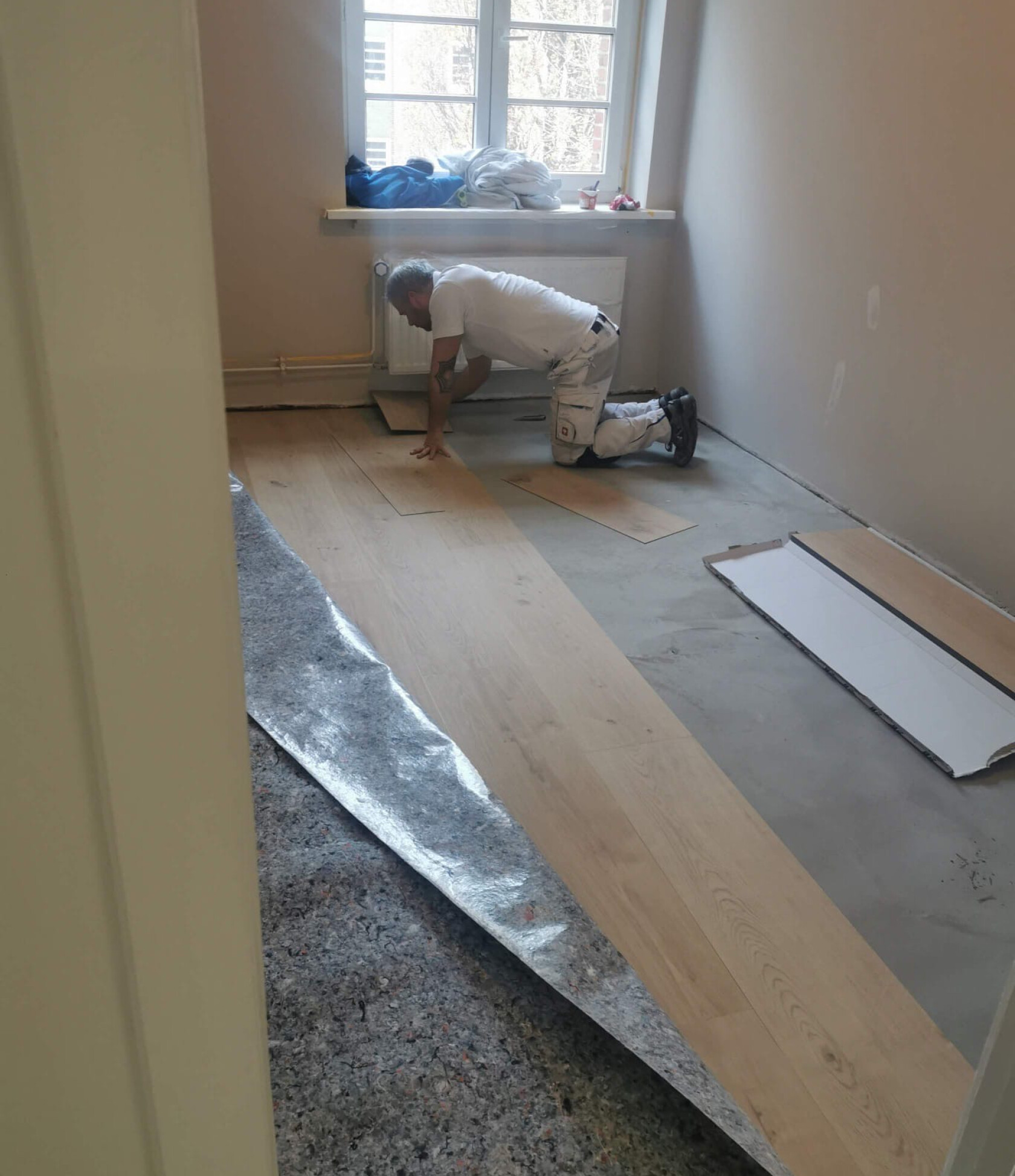 Mann kniet und verlegt Laminatpaneele im kleinen Raum; Unterlage teilweise aufgerollt, zugeschnittene Dielen und Kartonverpackung am Boden, Fenster mit Heizkörper im Hintergrund.