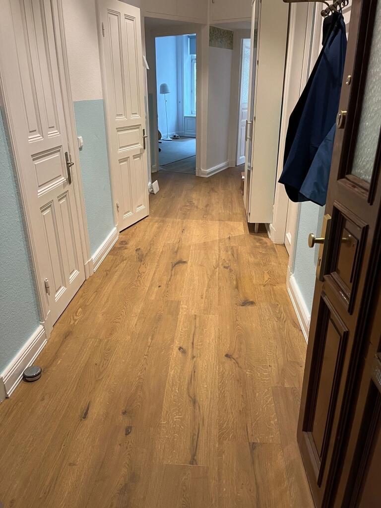 Breite Eichenholz-Dielen verlaufen durch einen Wohnungsflur, führen zu geöffneten weißen Türen und einem hellen Zimmer; rechts Garderobe mit Jacken, links Wandleisten und ein kleiner runder Saugroboter.