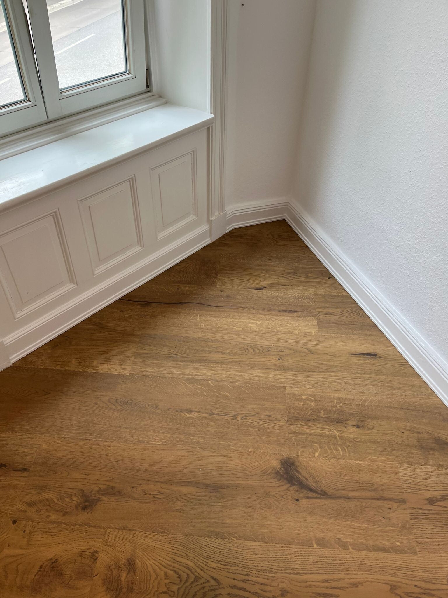 Eichenholz-Parkettboden mit sichtbarer Maserung, verlegt in Raumecke unter einer weißen, profilierten Fensterbrüstung mit Paneelen und Sockelleisten an glatten weißen Wänden.