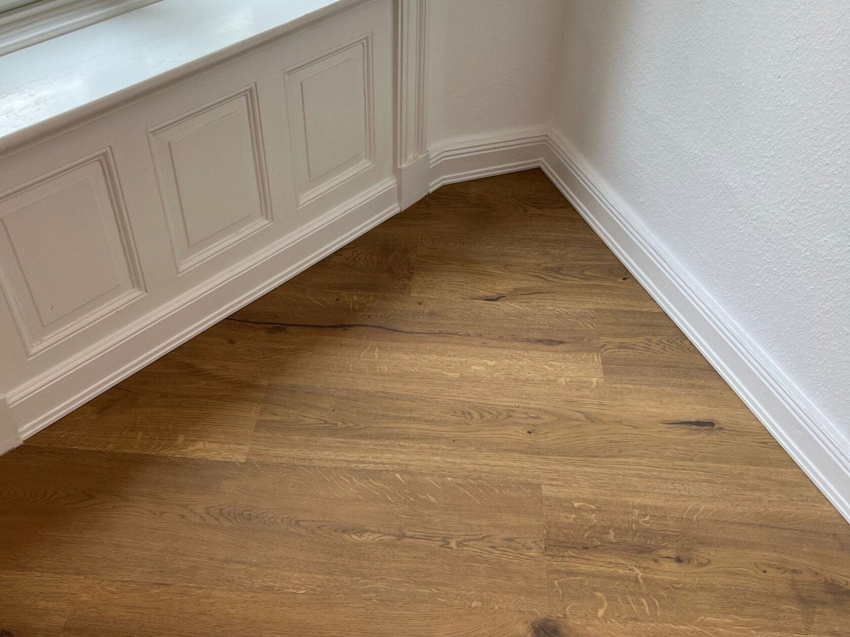 Eichenholz-Parkettboden mit sichtbarer Maserung, verlegt in Raumecke unter einer weißen, profilierten Fensterbrüstung mit Paneelen und Sockelleisten an glatten weißen Wänden.