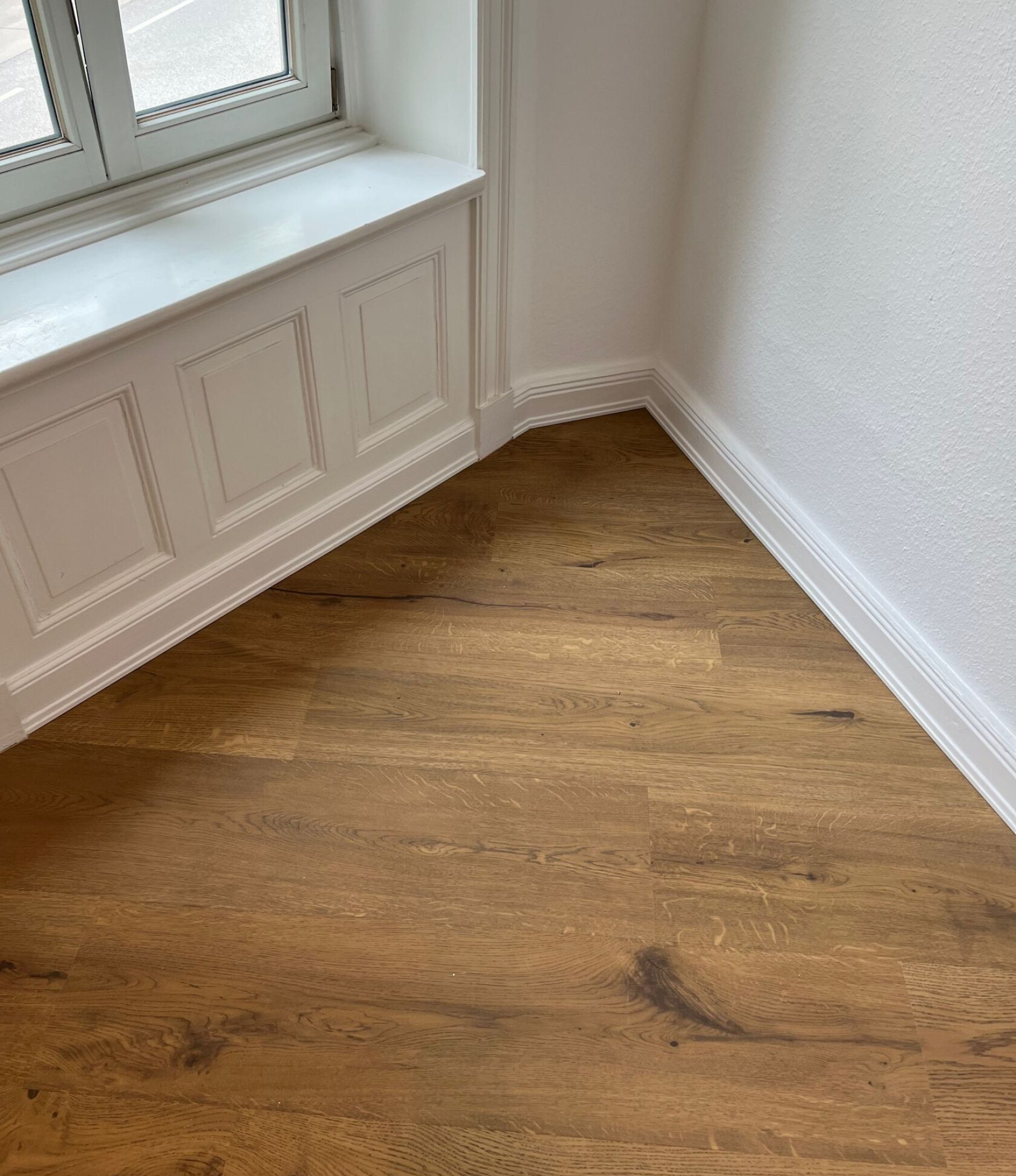 Eichenholz-Parkettboden mit sichtbarer Maserung, verlegt in Raumecke unter einer weißen, profilierten Fensterbrüstung mit Paneelen und Sockelleisten an glatten weißen Wänden.