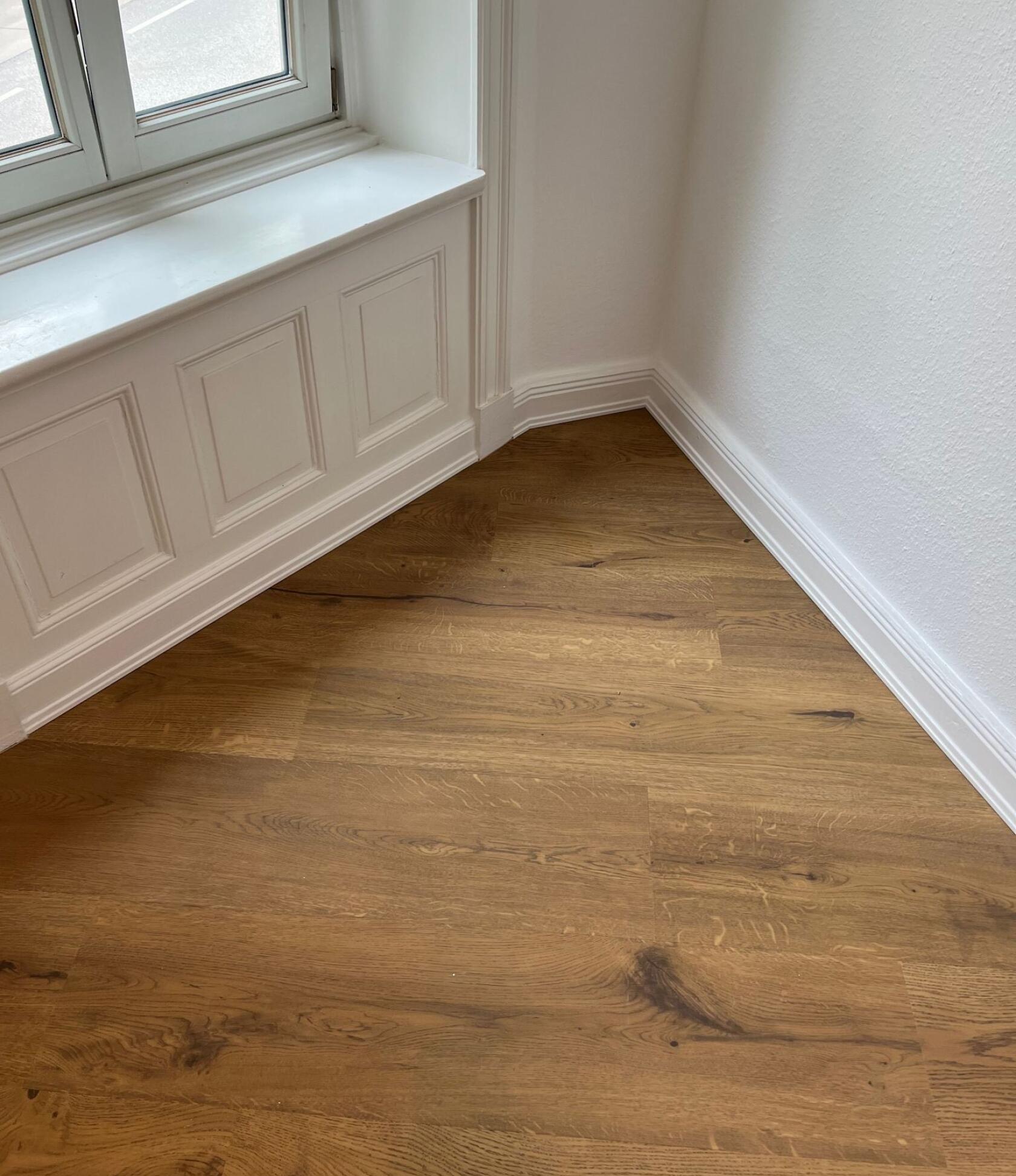 Eichenholz-Parkettboden mit sichtbarer Maserung, verlegt in Raumecke unter einer weißen, profilierten Fensterbrüstung mit Paneelen und Sockelleisten an glatten weißen Wänden.