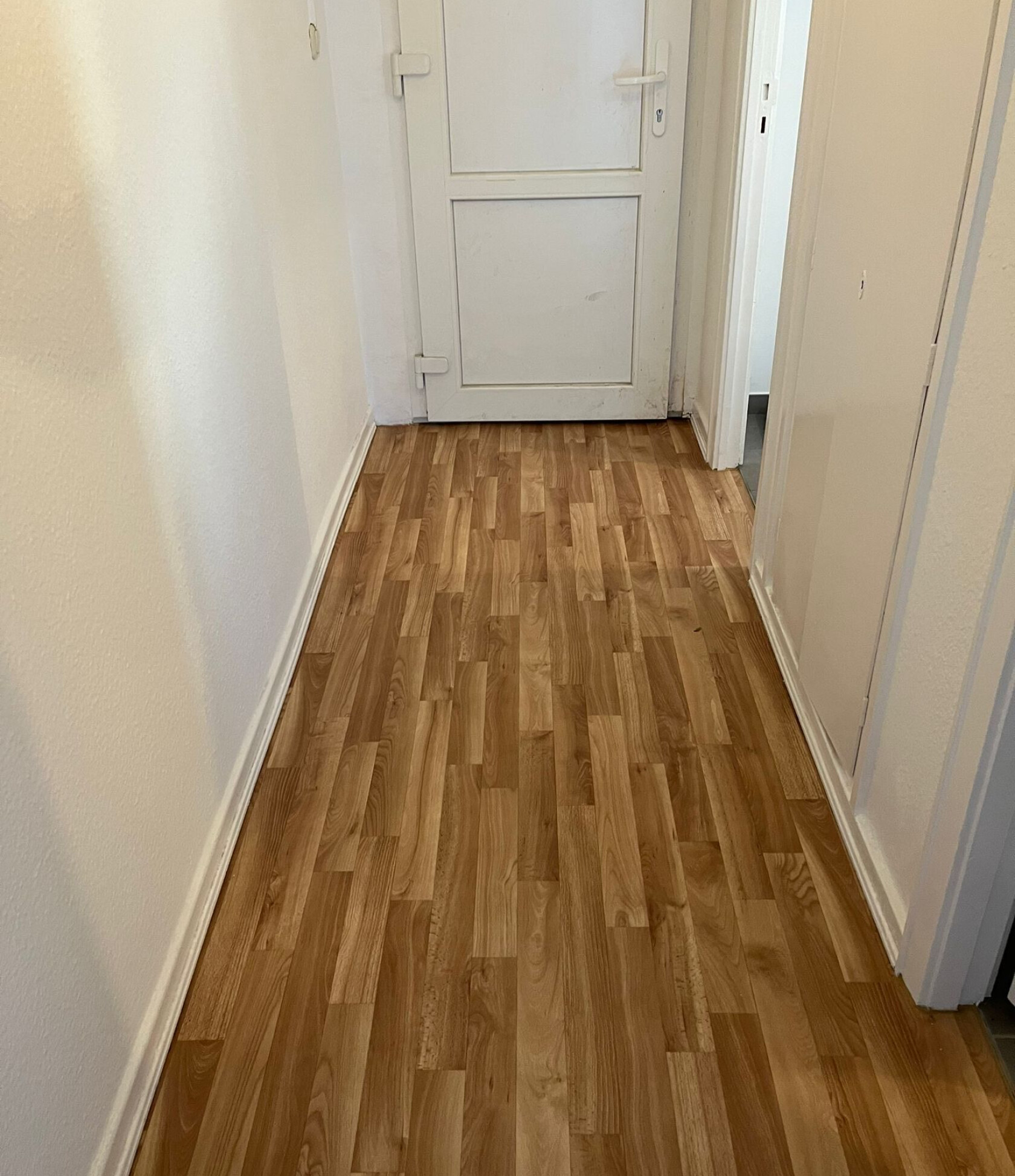 Flur mit hellbraunem Laminatboden – Boden verläuft zum Ende des Flurs – geschlossene weiße Tür, weiße Wände und Sockelleisten an beiden Seiten.