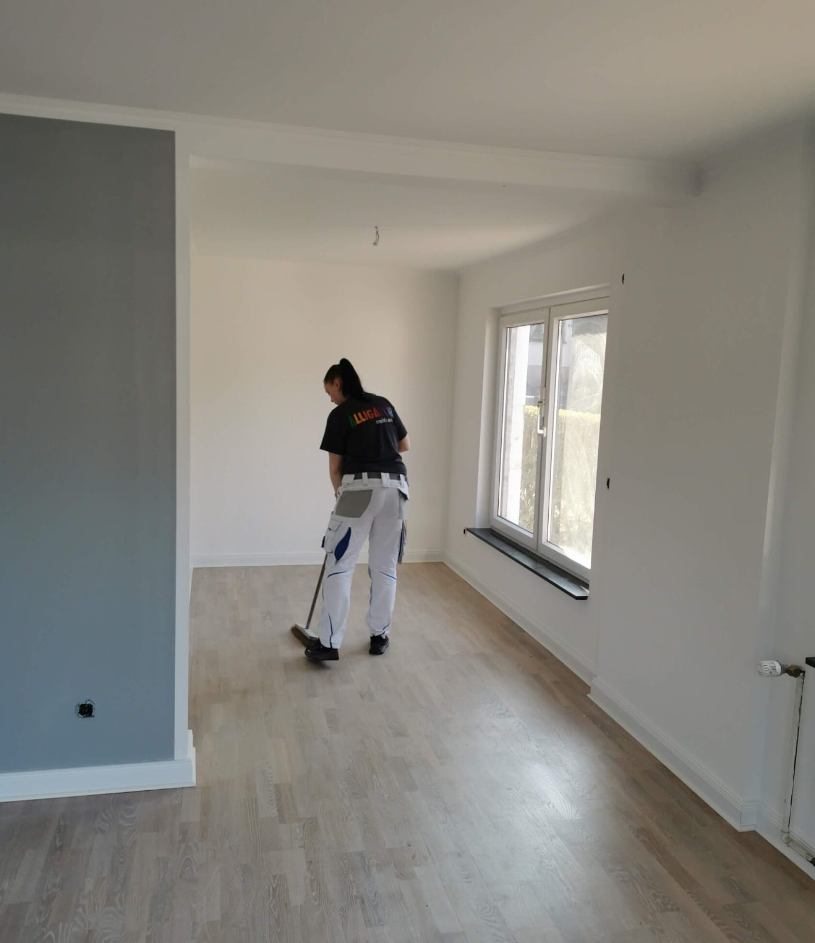 Person kehrt mit Besen hellen Holzboden in einem leeren, renovierten Raum mit weißen Wänden, großem Fenster rechts und grauer Akzentwand links.
