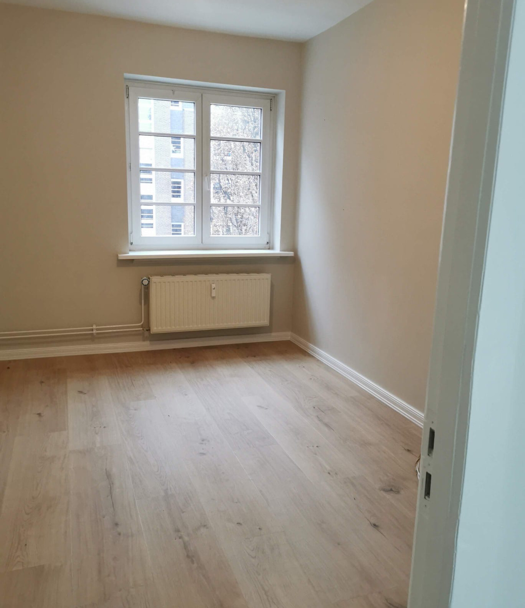Leeres Zimmer – ein weißgerahmtes Fenster mit Außenblick befindet sich über einem Wandheizkörper – heller Holzboden, hellbeige Wände und rechter Türrahmen sichtbar.