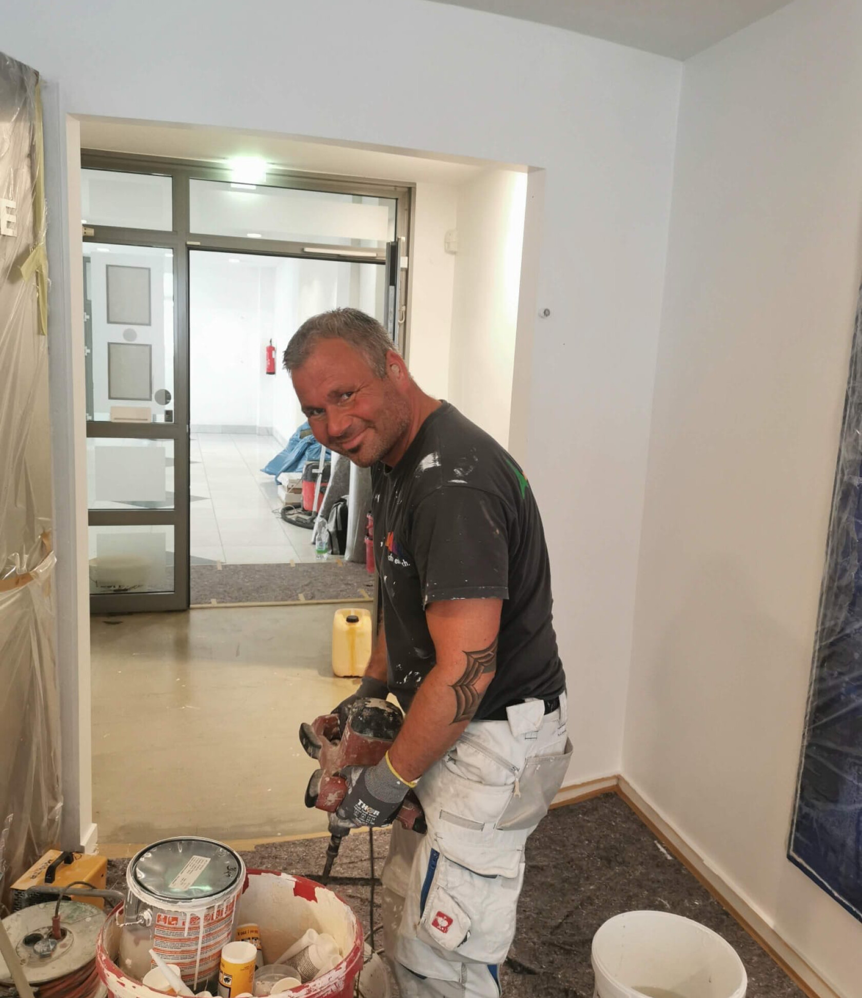 Handwerker bedient elektrischen Rührer über Eimer mit Farbe, steht in einem Innenraum mit Abdeckfolie und Schutzbelag, geöffnete Tür zum Flur im Hintergrund, Arbeitskleidung und Handschuhe.