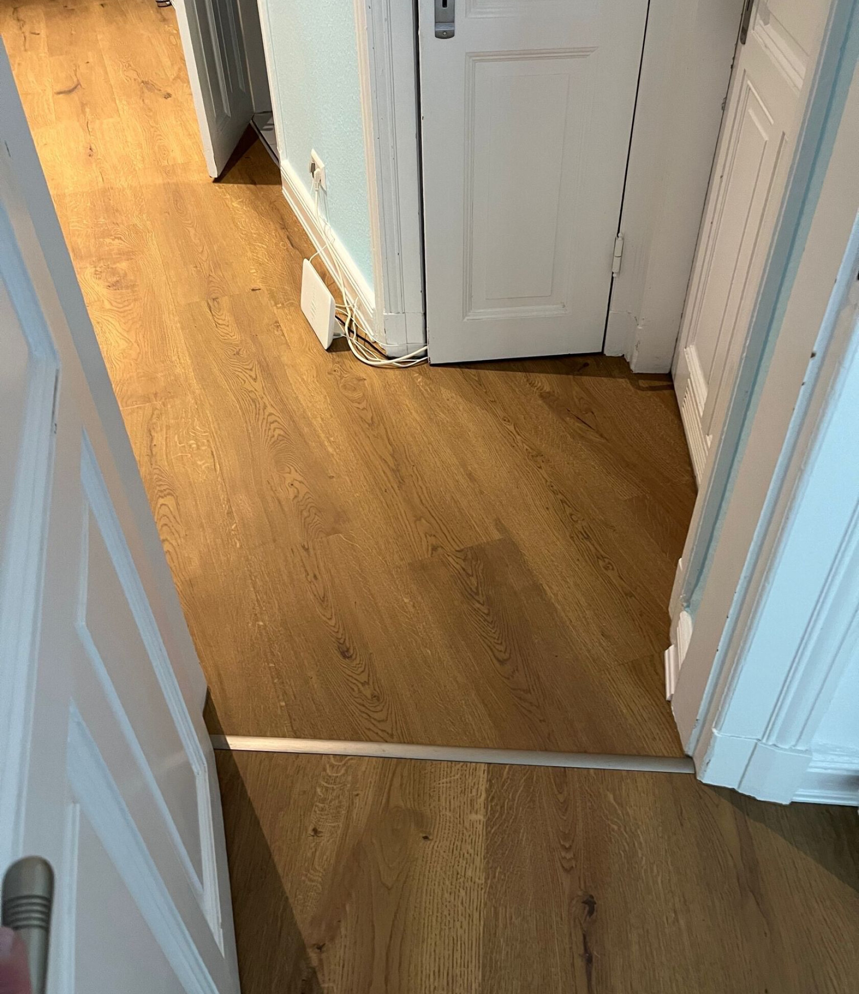 Holzfußboden mit metallischer Übergangsleiste, die eine Türöffnung überquert; weiße Tür und Türrahmen, an der Wand ein weißes Netzwerkgerät mit Kabeln am Sockel.