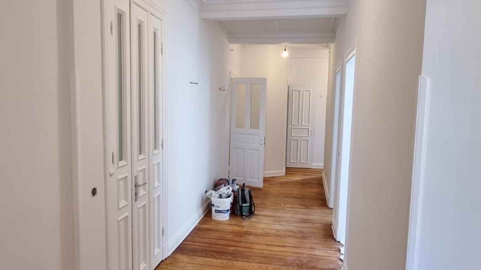 Schmaler weißer Flur – verläuft in die Tiefe – mit durchgehendem Holzdielenboden, mehreren weißen Türen, einer hängenden Glühbirne und Eimer sowie Werkzeugtasche am Boden.
