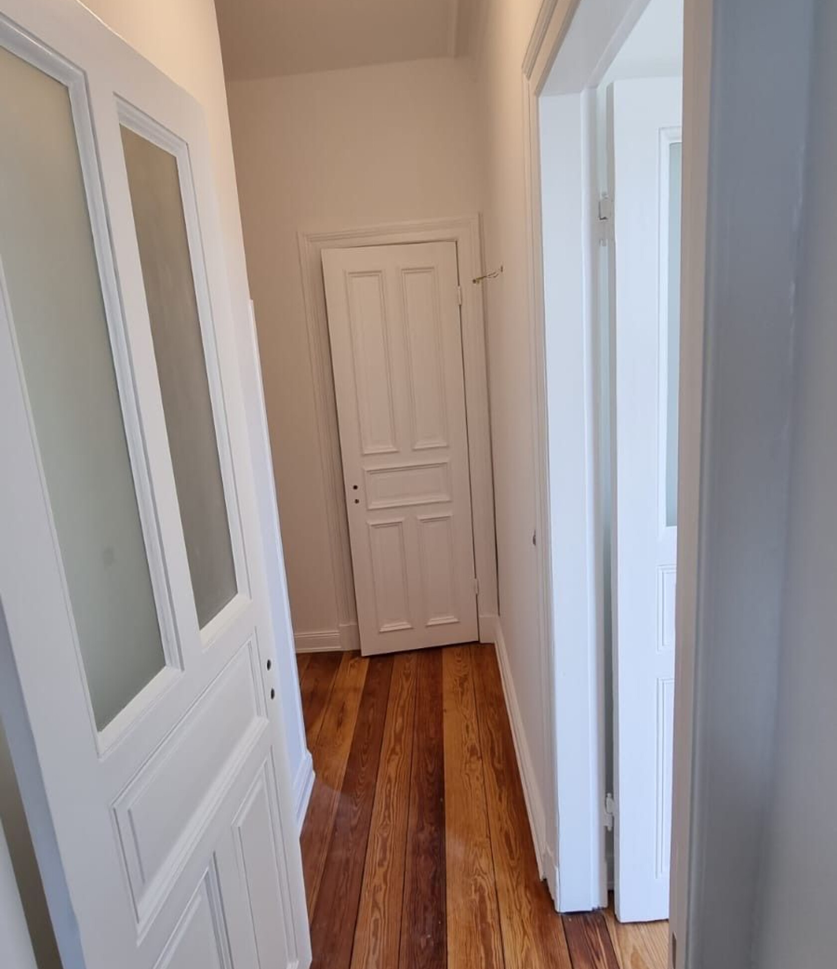 Schmale weiße Innentür am Ende eines engen Flurs — geschlossen; links geöffnete weiße Tür mit satiniertem Glas, rechts Türöffnung, helle Wände und dunkel-hell gestreifte Holzdielen.