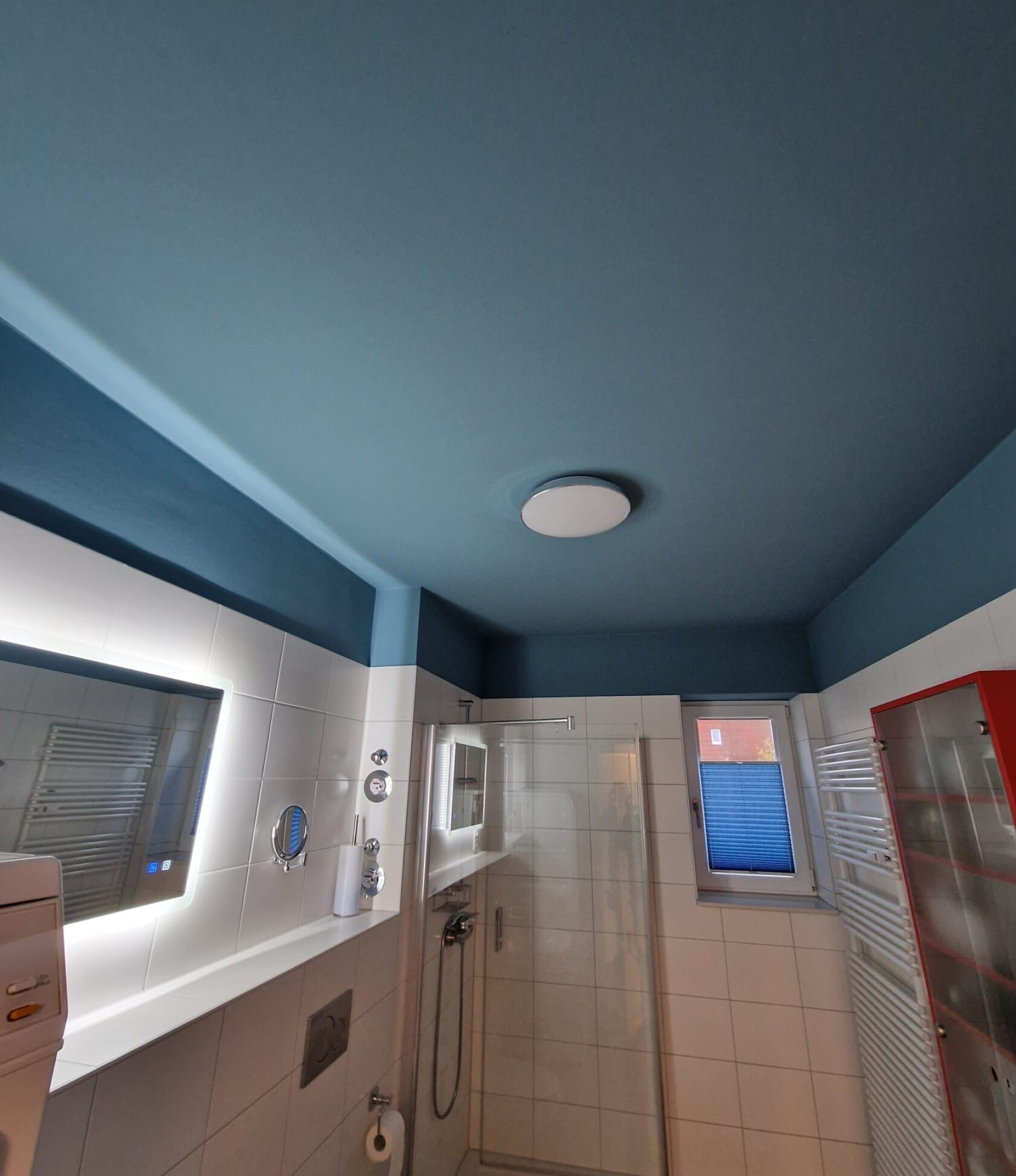 Blau gestrichene Badezimmerdecke mit runder Deckenleuchte über einer Glas-Duschkabine; weiße Fliesen, beleuchteter Wandspiegel, kleines Fenster mit blauem Plissee und Handtuchheizung.