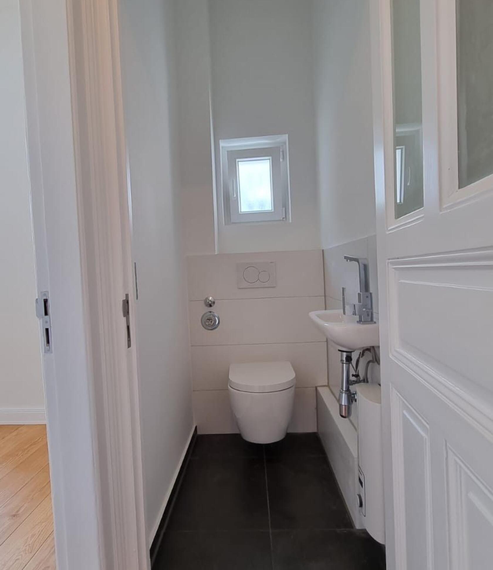Wandhängendes WC an der Rückwand montiert, in schmalem weißen Gästebad mit kleinem hochgesetztem Fenster, kleinem Waschbecken rechts und dunklem Fliesenboden.