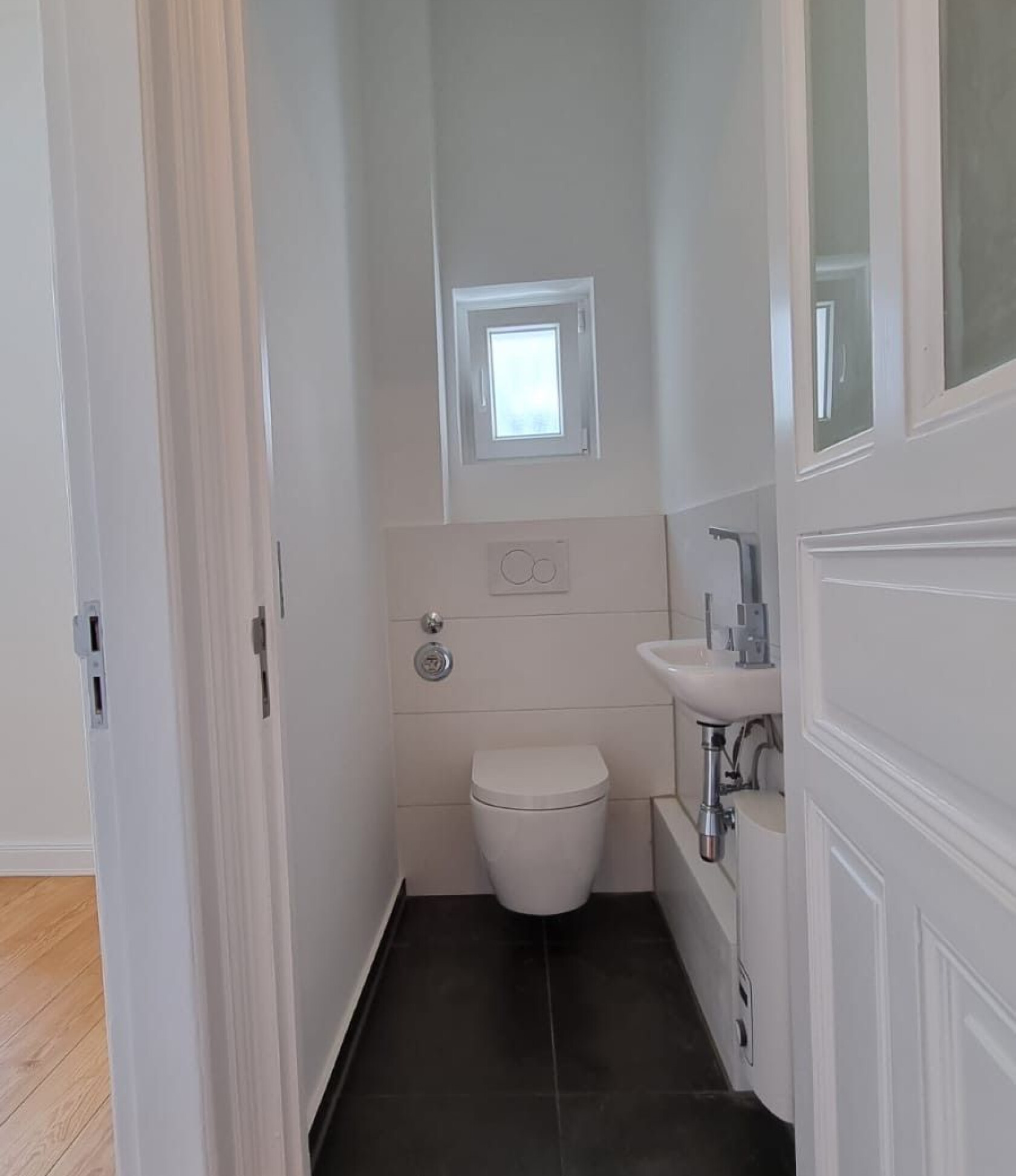 Wandhängendes WC an der Rückwand montiert, in schmalem weißen Gästebad mit kleinem hochgesetztem Fenster, kleinem Waschbecken rechts und dunklem Fliesenboden.