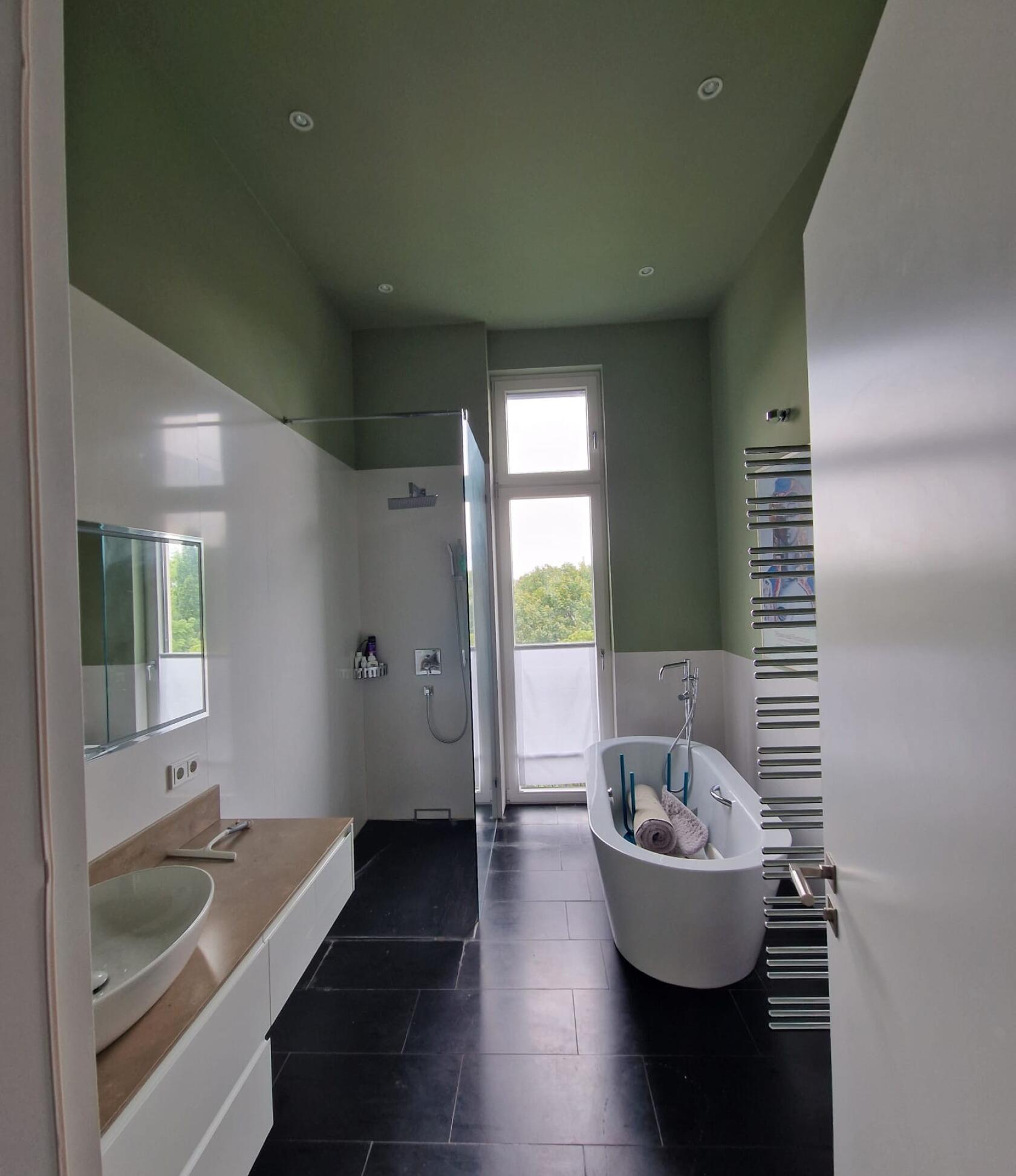 Freistehende weiße Badewanne steht rechts im Raum, enthält aufgerollte Handtücher und Badutensilien — links begehbare Dusche und Waschbecken, großes Fenster, dunkle Bodenfliesen, grün gestrichene Decke
