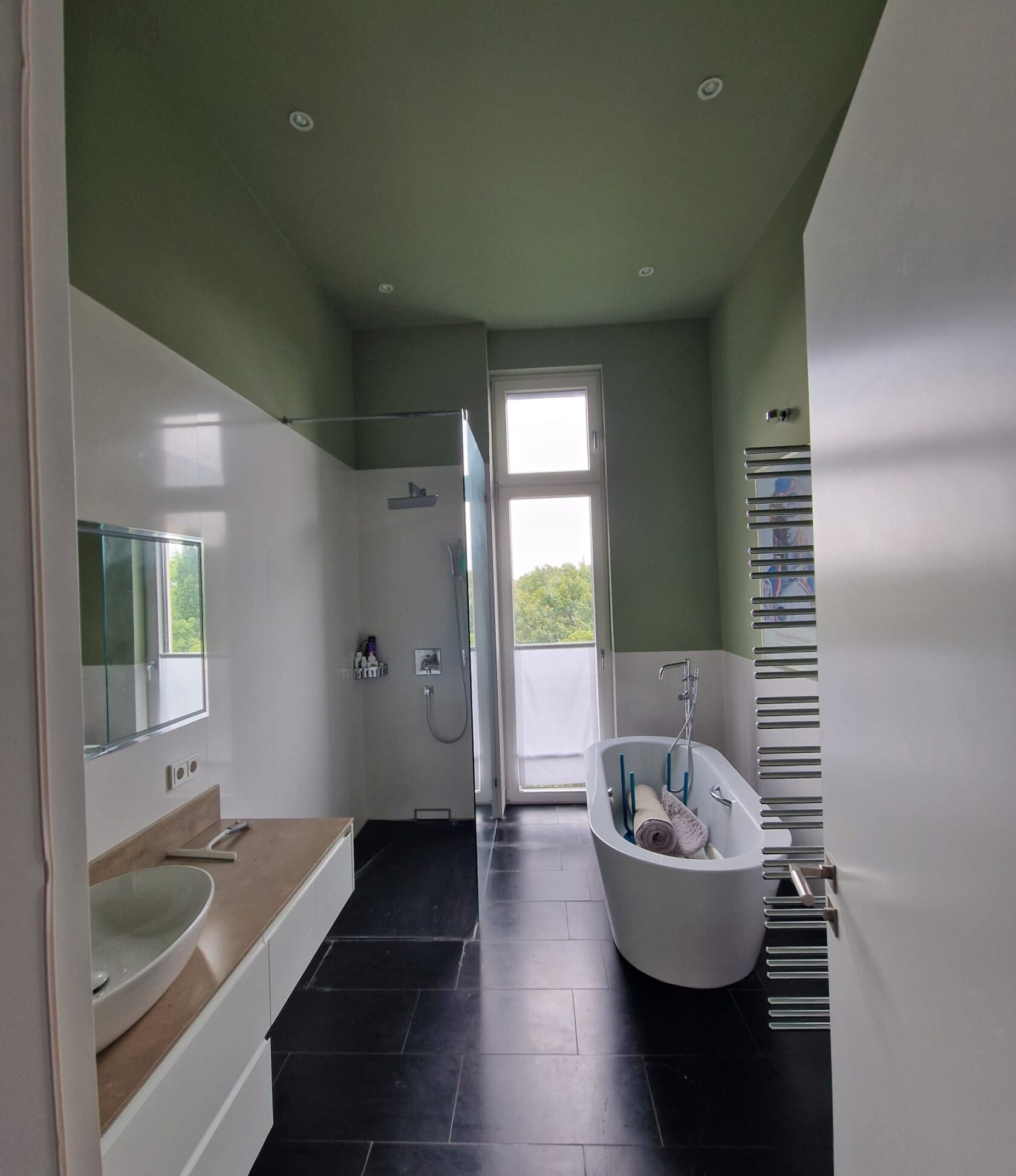 Freistehende weiße Badewanne steht rechts im Raum, enthält aufgerollte Handtücher und Badutensilien — links begehbare Dusche und Waschbecken, großes Fenster, dunkle Bodenfliesen, grün gestrichene Decke