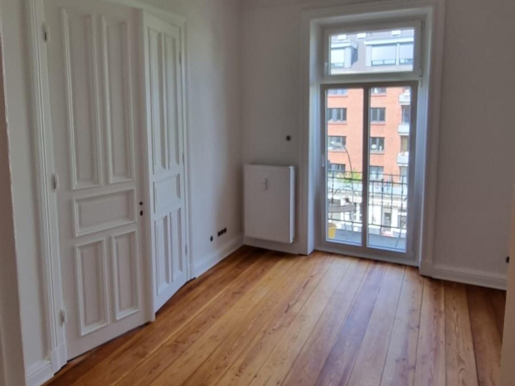 Leeres Zimmer mit breiten Holzdielen, geschlossener weißer Innentür links und verglaster Balkontür rechts, die auf eine gegenüberliegende Backsteinfassade hinausblickt.