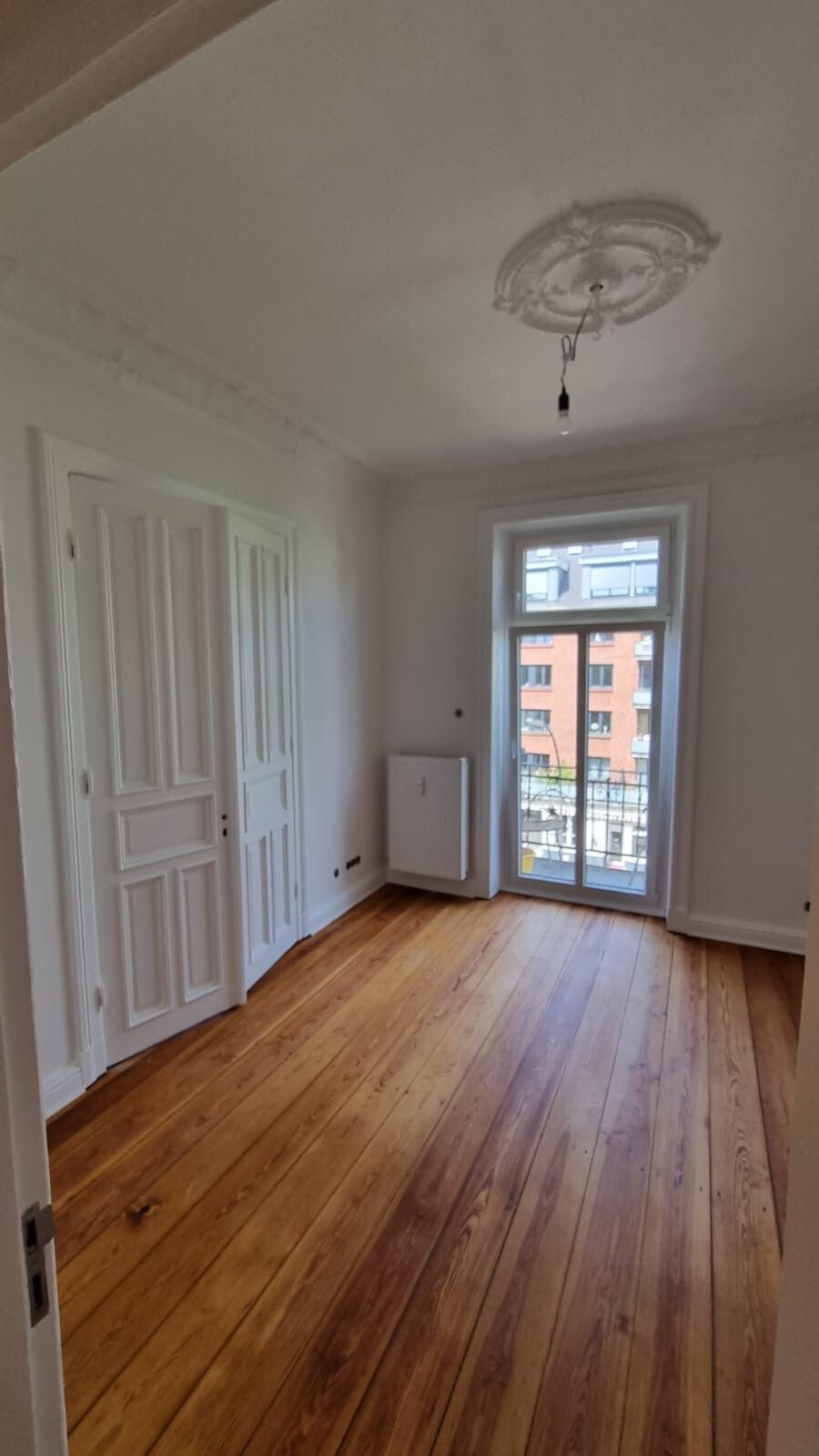 Leeres Zimmer mit breiten Holzdielen, geschlossener weißer Innentür links und verglaster Balkontür rechts, die auf eine gegenüberliegende Backsteinfassade hinausblickt.