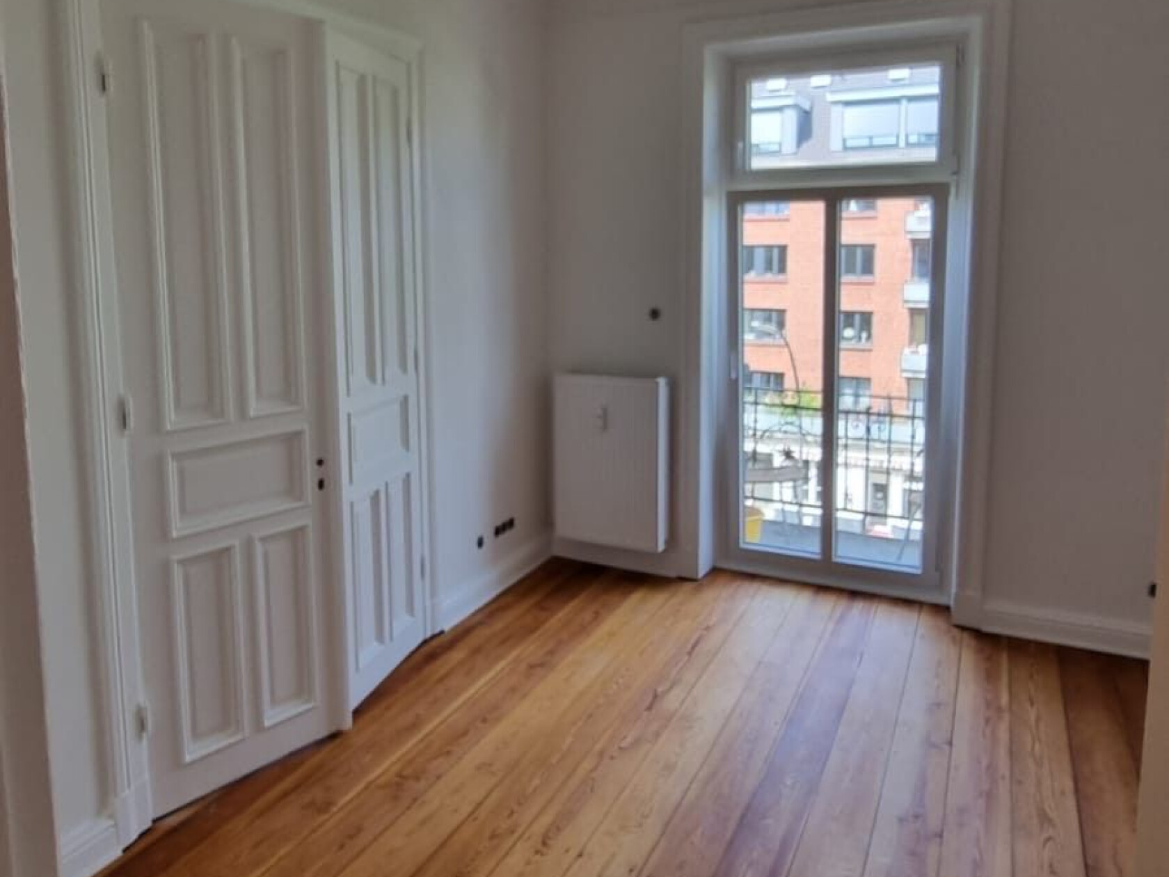 Leeres Zimmer mit breiten Holzdielen, geschlossener weißer Innentür links und verglaster Balkontür rechts, die auf eine gegenüberliegende Backsteinfassade hinausblickt.