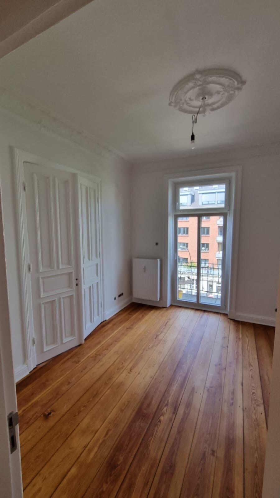 Leeres Zimmer mit breiten Holzdielen, geschlossener weißer Innentür links und verglaster Balkontür rechts, die auf eine gegenüberliegende Backsteinfassade hinausblickt.