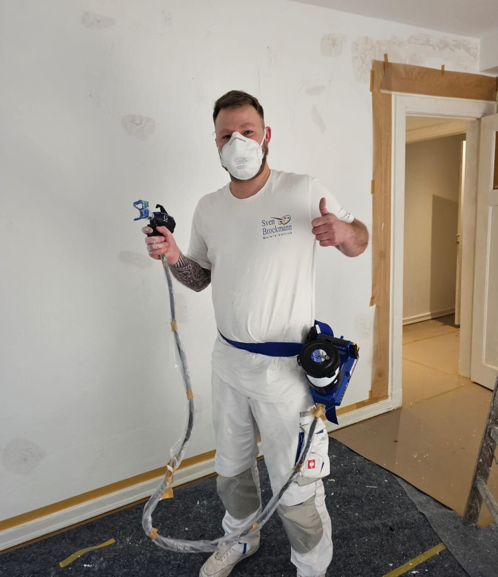 Person in weißer Arbeitskleidung mit Schutzmaske, Logo 'Sven Brockmann Malerbetrieb' auf dem T‑Shirt — hält Farbspritzpistole mit Schlauch an gurtgetragenem Gerät, zeigt Daumen hoch; Innenraum mit abgeklebtem Türrahmen und abgedecktem Boden.