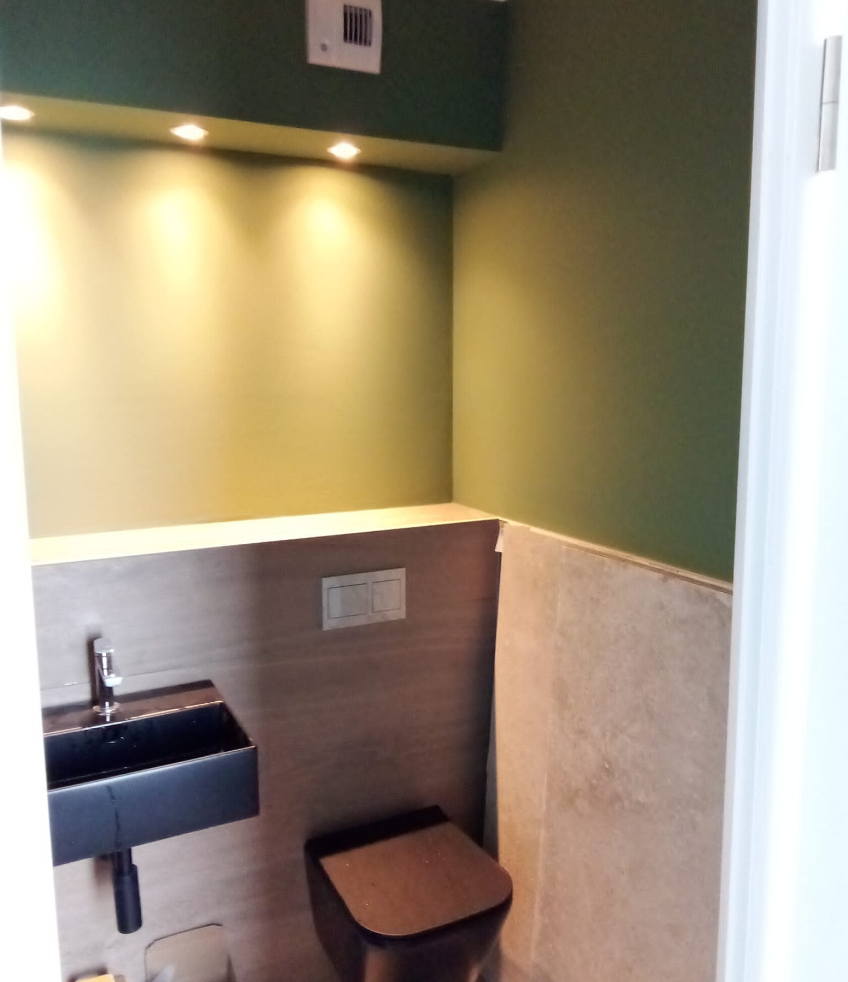 Schwarze wandhängende Toilette und kleines schwarzes Waschbecken, an der Wand montiert; schmale Ablage darüber, grüne Wände mit drei eingelassenen Strahlern und halbhoher heller Wandverkleidung.