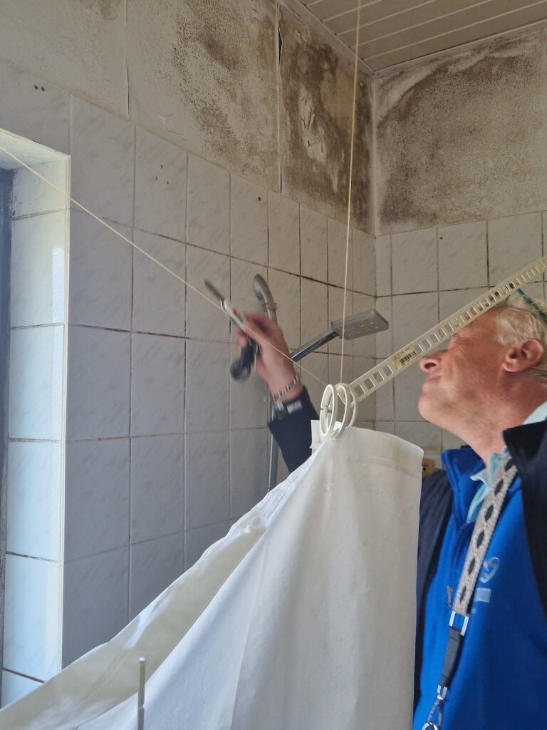 Person zieht Wäscheklammern an einer aufgehängten Leine im kleinen gefliesten Badezimmer über der Dusche; Duschkopf sichtbar, ausgeprägte schwarze Schimmelstellen an Decke und Wand.