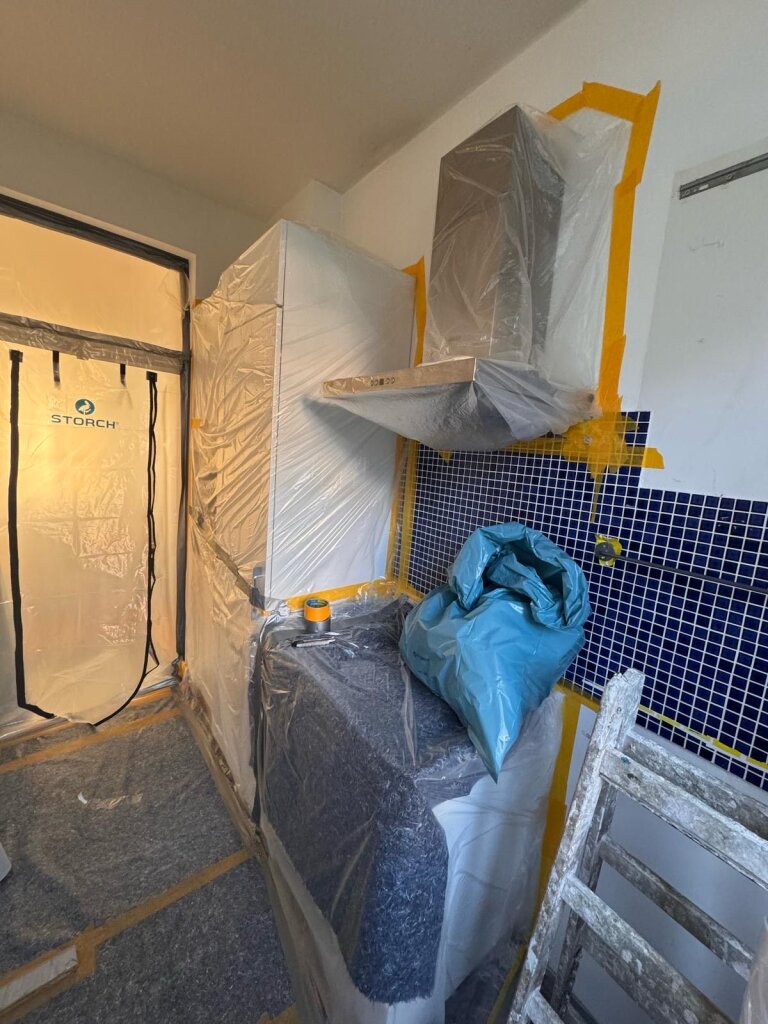 Küchenzeile, Schränke und Geräte mit Kunststofffolie und gelbem Malerkrepp abgedeckt, Dunstabzugshaube verhüllt, blauer Mosaikfliesen‑Fliesenspiegel, blaue Abdeckplane auf Arbeitsfläche, Staubschutztür mit Schriftzug 